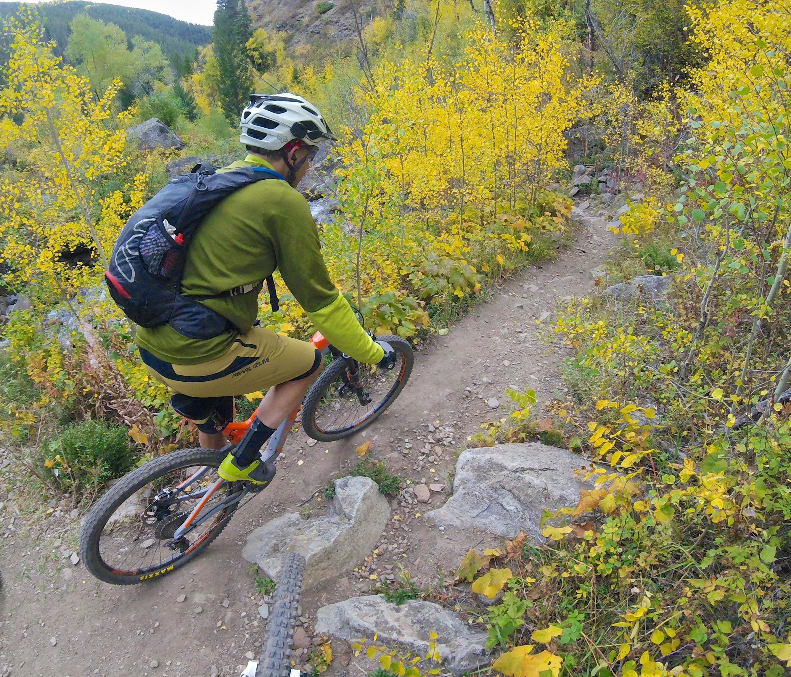 A person riding a mountain bike on a dirt trail surrounded by vibrant yellow foliage and greenery. The cyclist, wearing a helmet and a green long-sleeve shirt with shorts, navigates a rocky path in a wooded area during autumn. Two Elk via Vail Pass mountain bike trail.