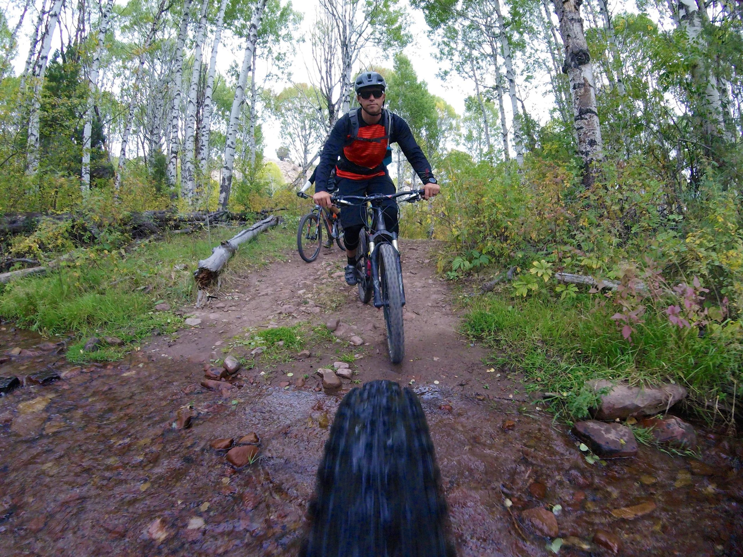 Two mountain bikers ride along a dirt trail surrounded by lush greenery and birch trees. The foreground shows a close-up of a bike tire approaching a small stream, while one rider is visible in the mid-ground, wearing sunglasses and a helmet, with a colorful backpack. The scene captures the essence of outdoor adventure and biking in nature. Hardscrabble Mountain Trails mountain bike trail.