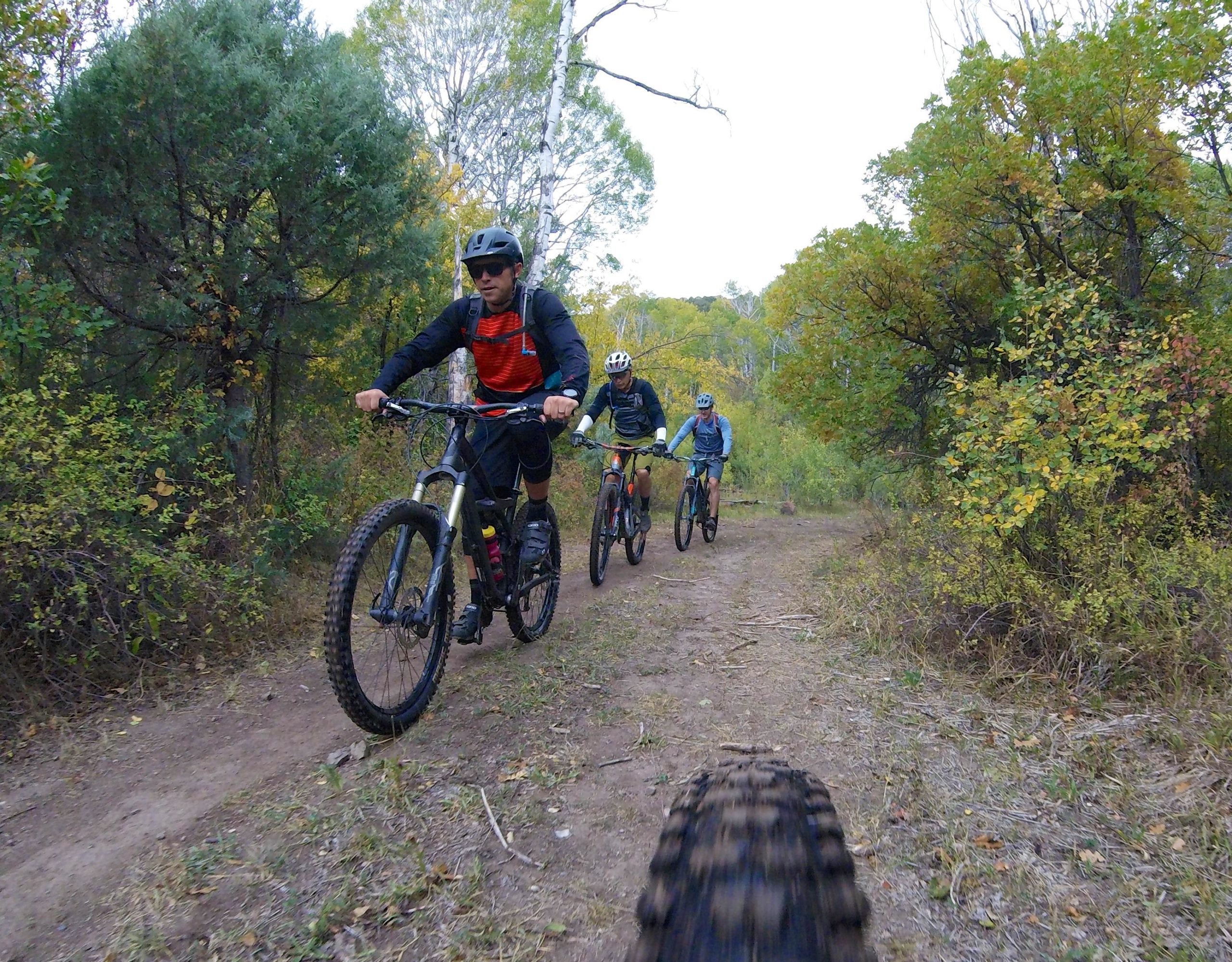 Three mountain bikers riding along a dirt trail in a lush, green forest during autumn. The first rider, wearing a helmet and a red and black jersey, leads the group, followed by two others in blue and yellow gear. The scene captures the vibrant foliage in the background, showcasing seasonal colors. Hardscrabble Mountain Trails mountain bike trail.
