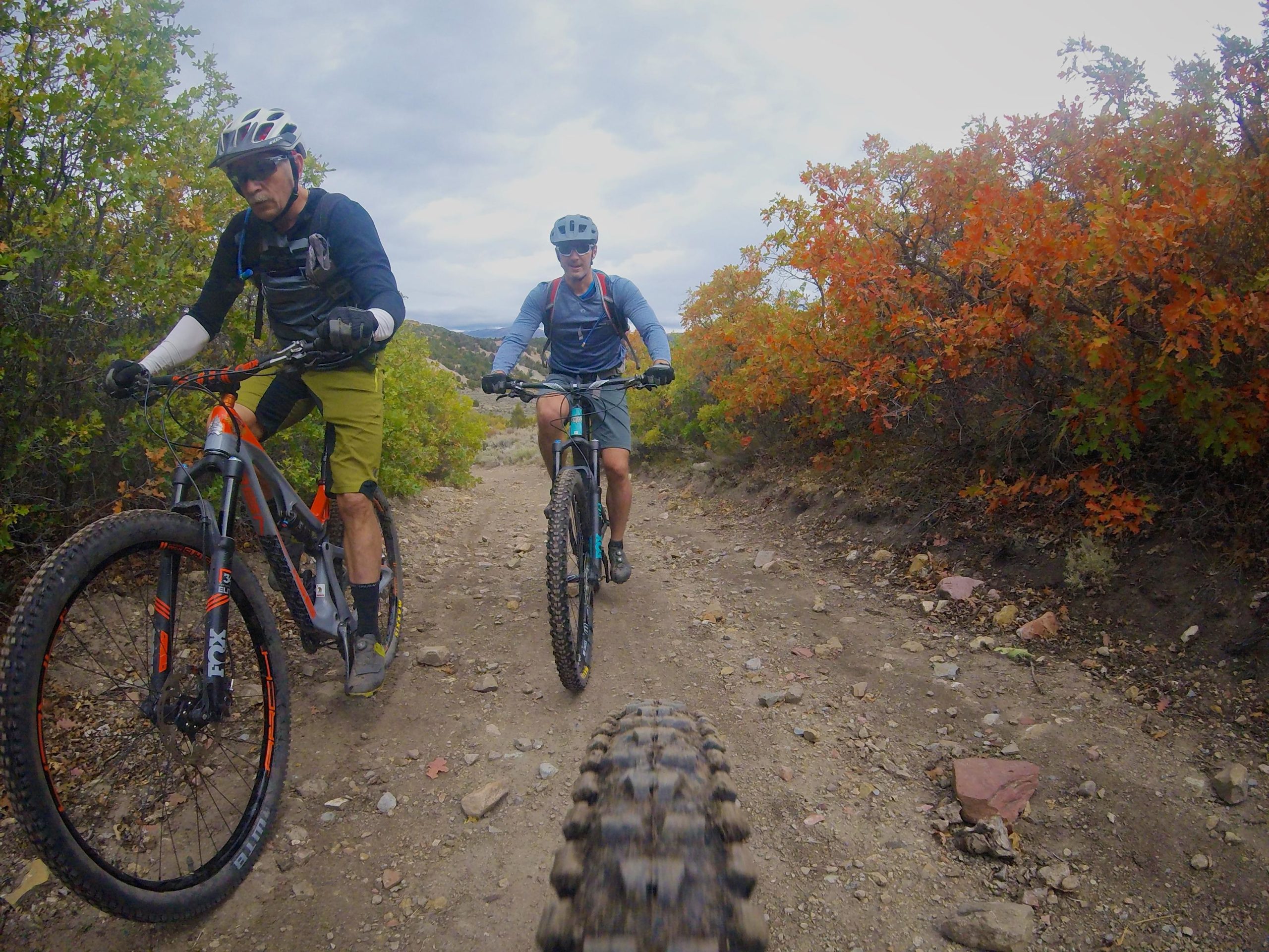 Two mountain bikers ride along a gravel path surrounded by vibrant fall foliage. The first rider is in the foreground, wearing a black long-sleeve shirt and green shorts, while the second rider is slightly behind, dressed in a teal shirt and gray shorts. Both cyclists are focused on the trail ahead, with rocky terrain and colorful bushes visible on either side. Hardscrabble Mountain Trails mountain bike trail.