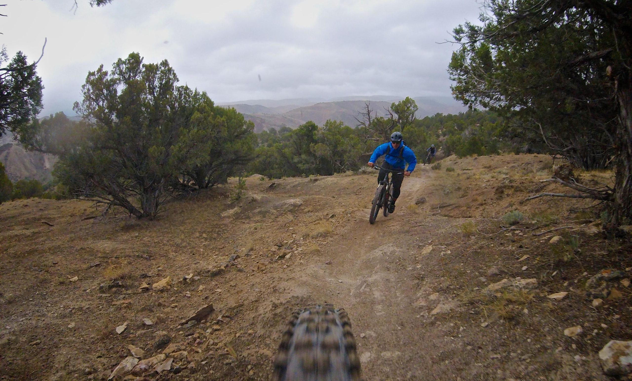 A mountain biker in a blue jacket navigates a rocky trail with trees on both sides, while another cyclist follows in the background. The cloudy sky hints at potential rain, creating a dynamic outdoor atmosphere. Hardscrabble Mountain Trails mountain bike trail.