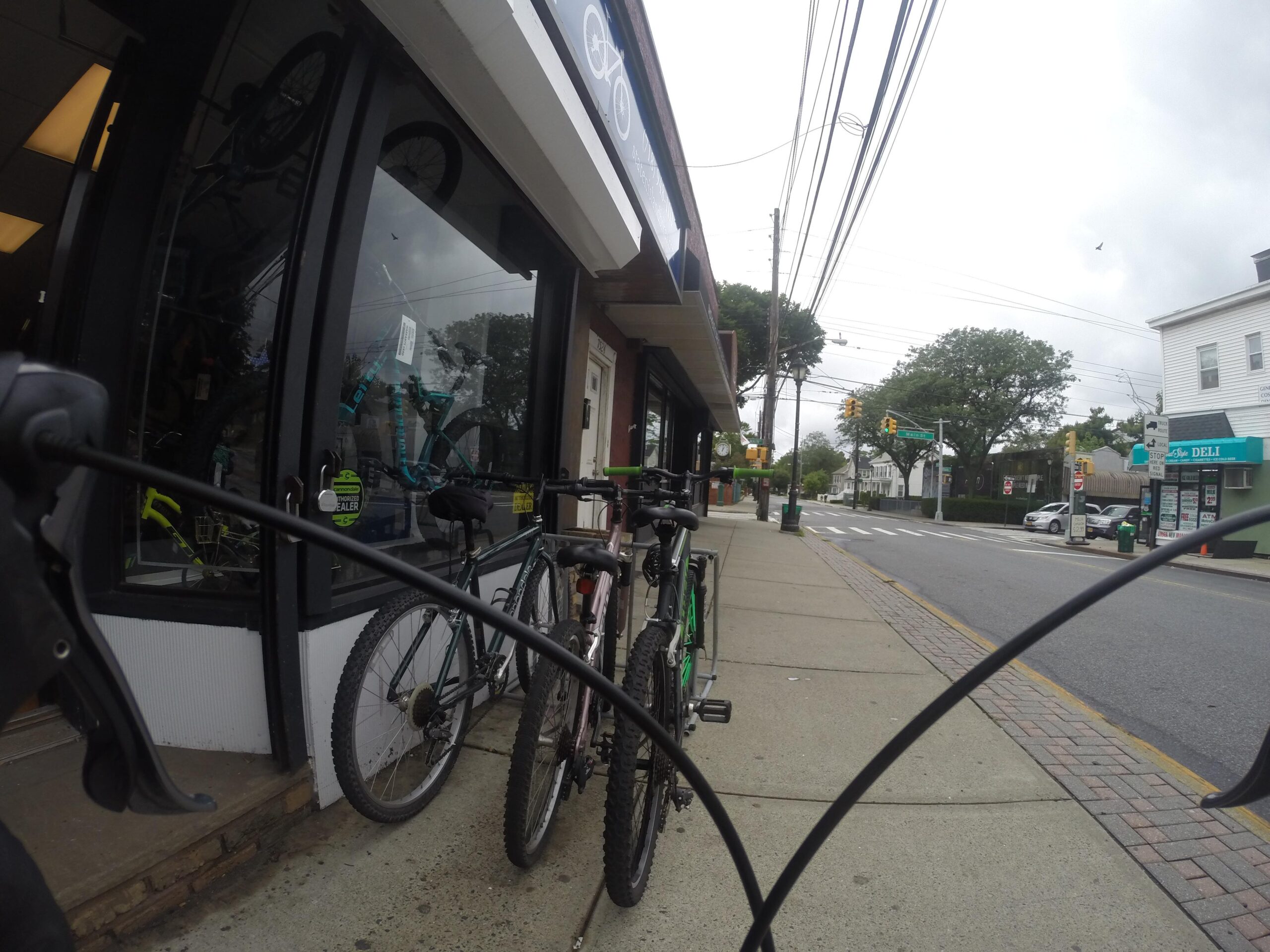 A view from a bicycle parked outside a bike shop, showing several bikes along the sidewalk. The shop's glass window displays more bicycles. The street is lined with utility poles and trees, with a deli visible in the background. The sky is overcast, indicating a cloudy day.