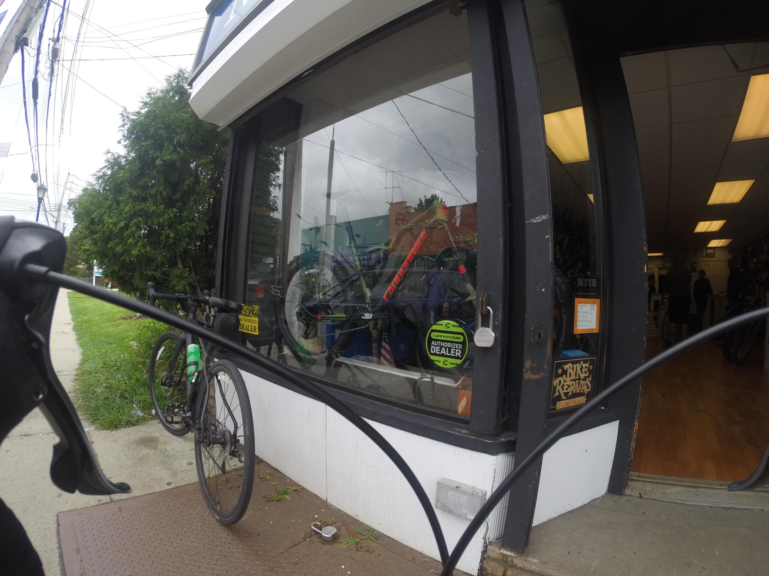 A view of a bicycle shop from the sidewalk, featuring a large window displaying various bicycles inside. The shop has a sign indicating it offers bike repairs. In the foreground, a bicycle is parked near the entrance. The scene is partially obscured by the handlebars of the parked bike, with greenery visible in the background.