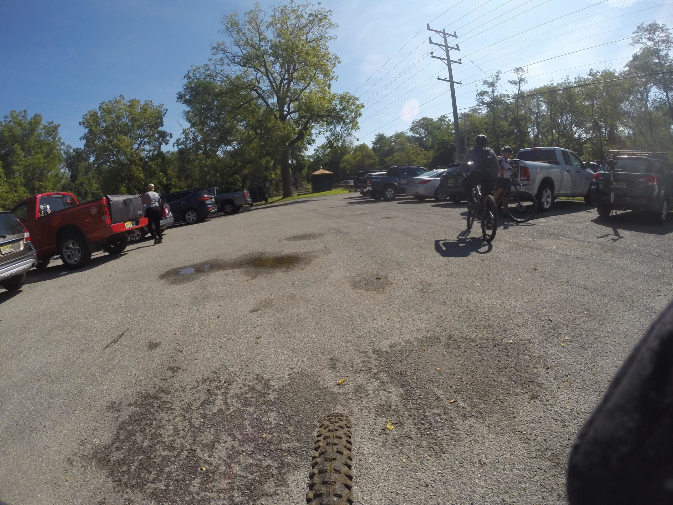 A sunny day in a parking lot with various vehicles parked, including a red truck and several SUVs. In the foreground, a biker is about to ride away, while another person is seen in the background near the truck. Trees line the edge of the parking area, and power lines are visible in the sky. Allaire State Park mountain bike trail.