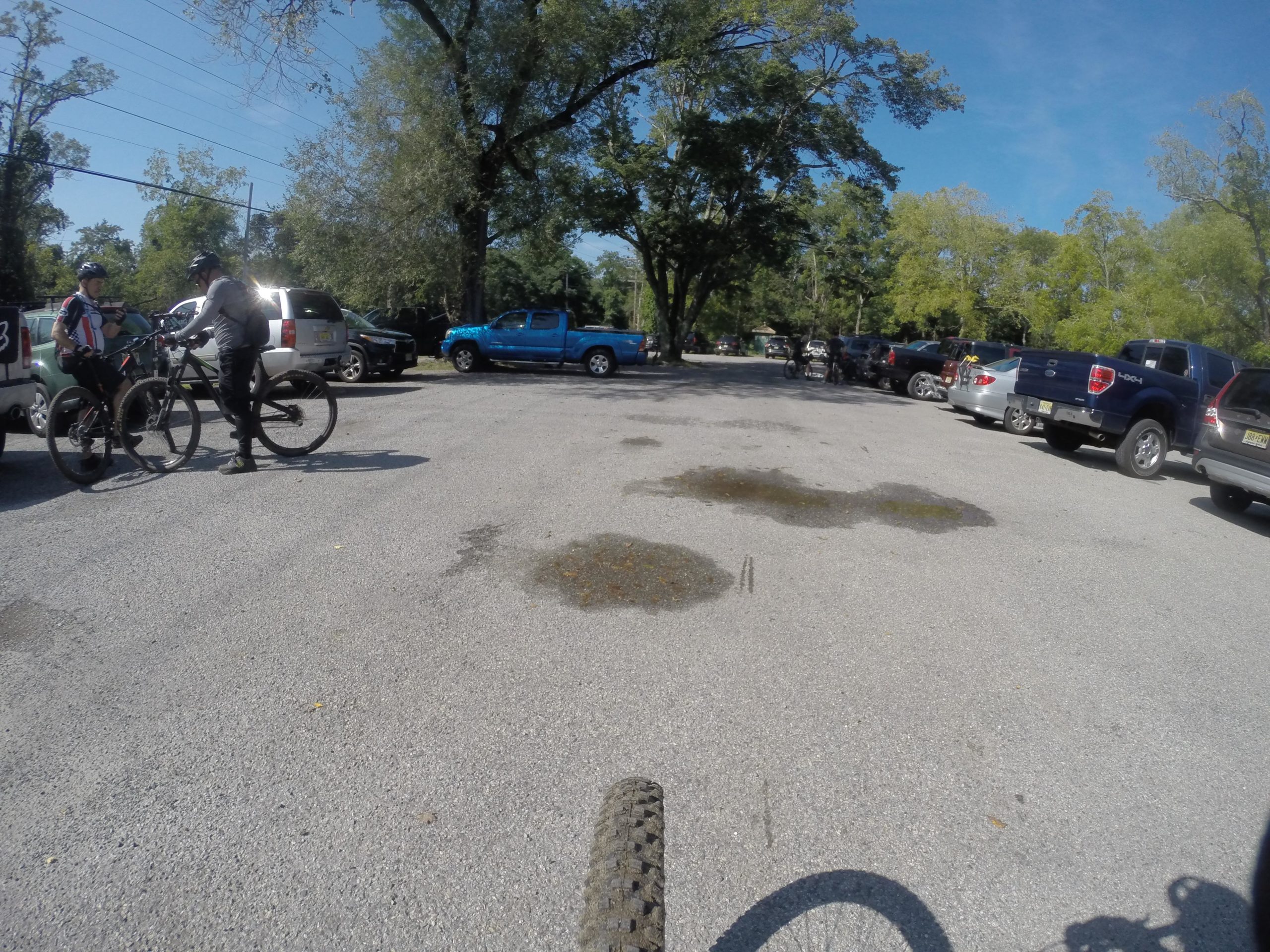 A view of a parking lot with several cars, featuring two cyclists engaged in conversation near their bikes. The scene is set on a sunny day, with trees in the background and a clear blue sky above. Puddles of water can be seen on the pavement. Allaire State Park mountain bike trail.