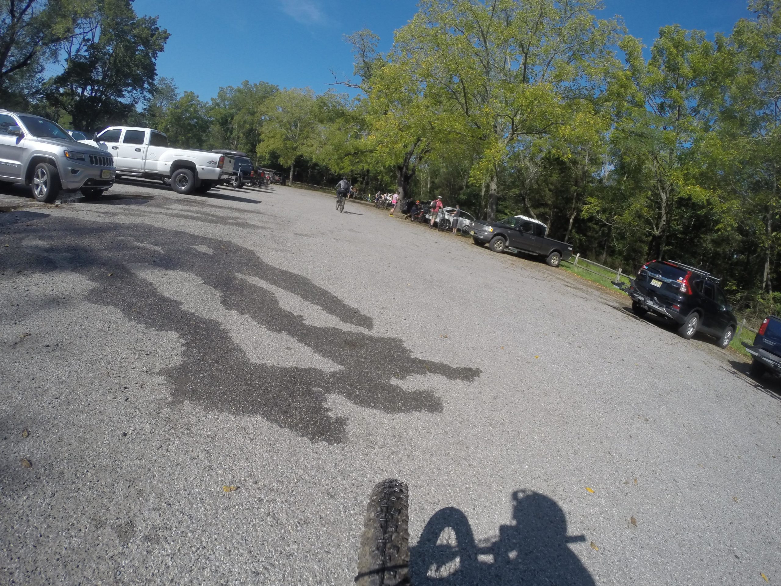 A gravel parking lot filled with various parked vehicles, including trucks and SUVs. In the foreground, the shadow of a bike is visible, suggesting a cyclist in motion. Trees with green leaves line the background, and a group of people can be seen near the edge of the parking area, indicating a recreational setting on a sunny day. Allaire State Park mountain bike trail.
