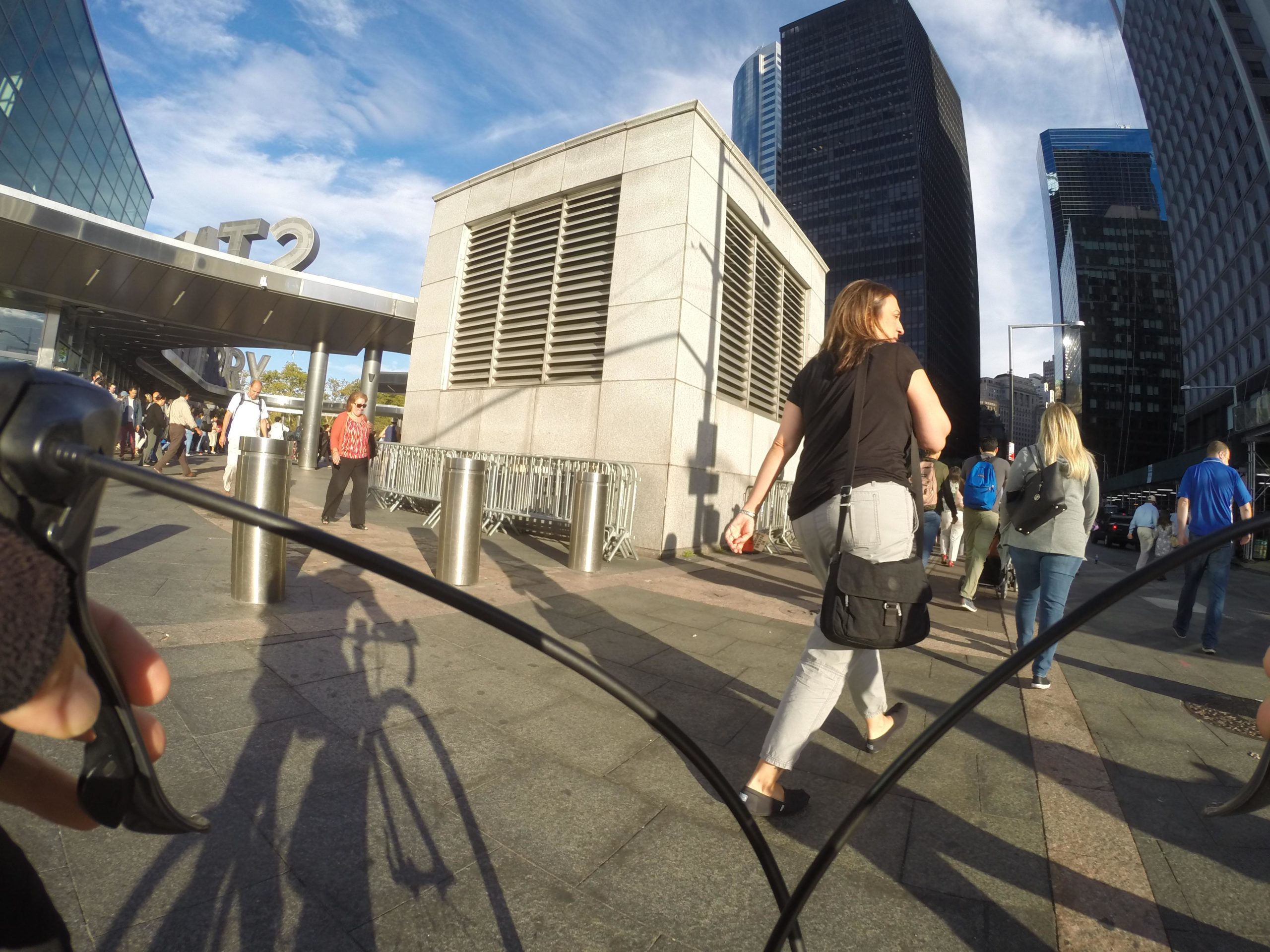 Image showing a city street scene with pedestrians walking past modern buildings. In the foreground, a person's hand is holding a bicycle handlebar, partially visible, casting a shadow on the ground. The background features other people, some wearing casual clothing, and a prominent sign with the letters "MT2." The sky above is partly cloudy. West Street Greenway mountain bike trail.