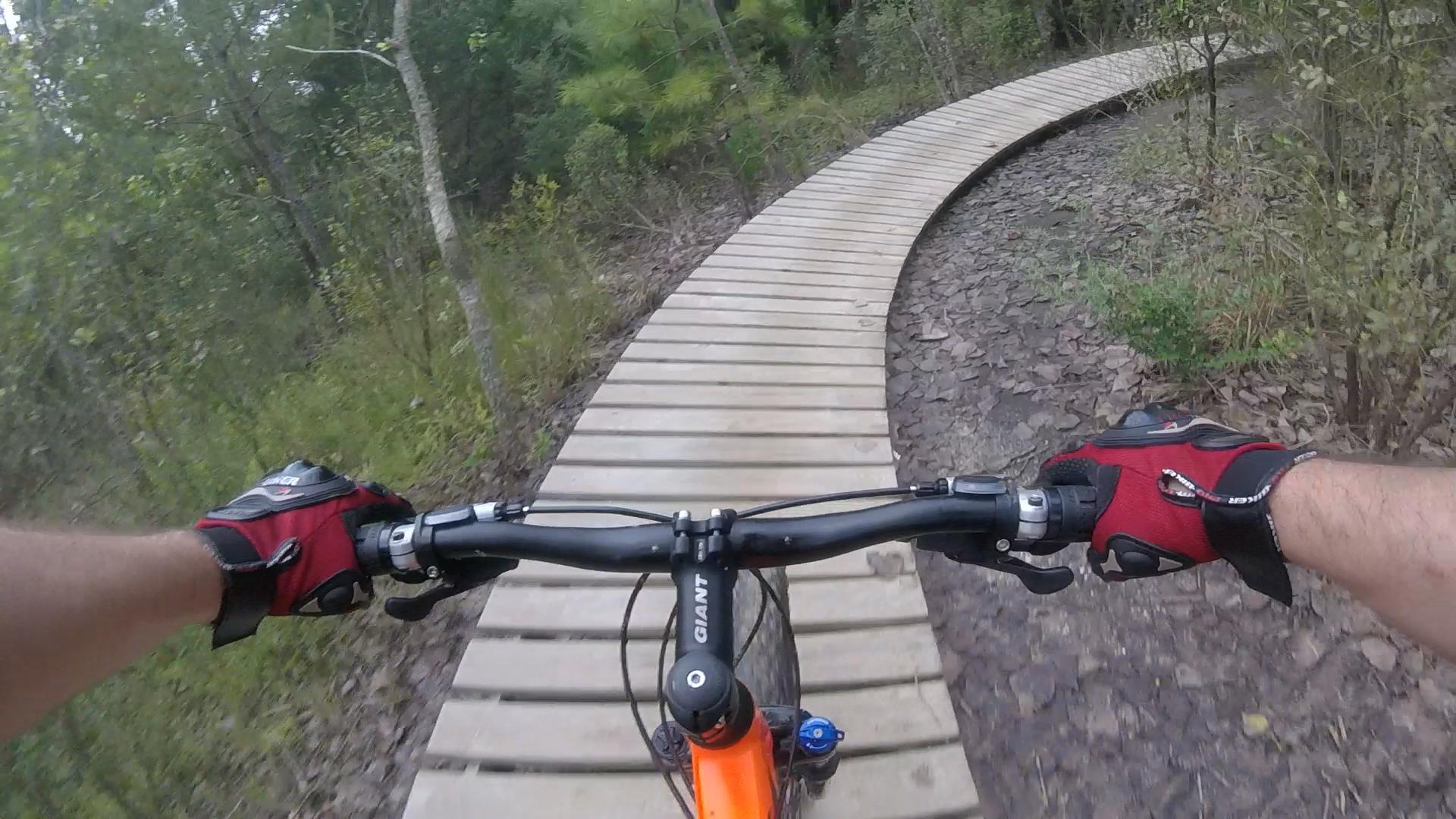 A person's hands gripping the handlebars of a mountain bike, viewed from the rider's perspective as they navigate a curved wooden pathway through a lush forest. The path is surrounded by trees and rocky terrain. Horry County Bike Run Park mountain bike trail.