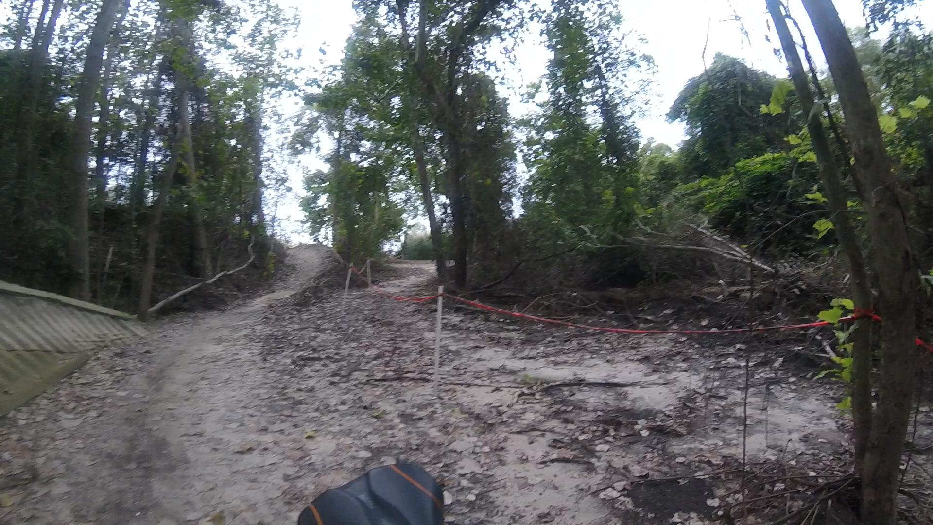 A narrow dirt path winding through a wooded area, flanked by trees and scattered leaves. A hill rises in the distance, marked with red caution tape. Horry County Bike Run Park mountain bike trail.