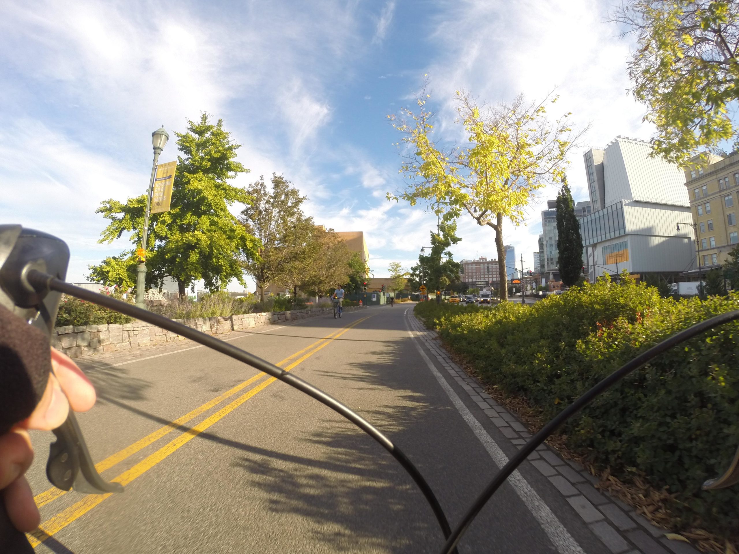 A cyclist's perspective on a wide path lined with trees, featuring green foliage and a clear blue sky. In the background, a modern building and signage can be seen along with another cyclist in motion. The image captures the feeling of an outdoor riding experience in an urban environment. West Street Greenway mountain bike trail.