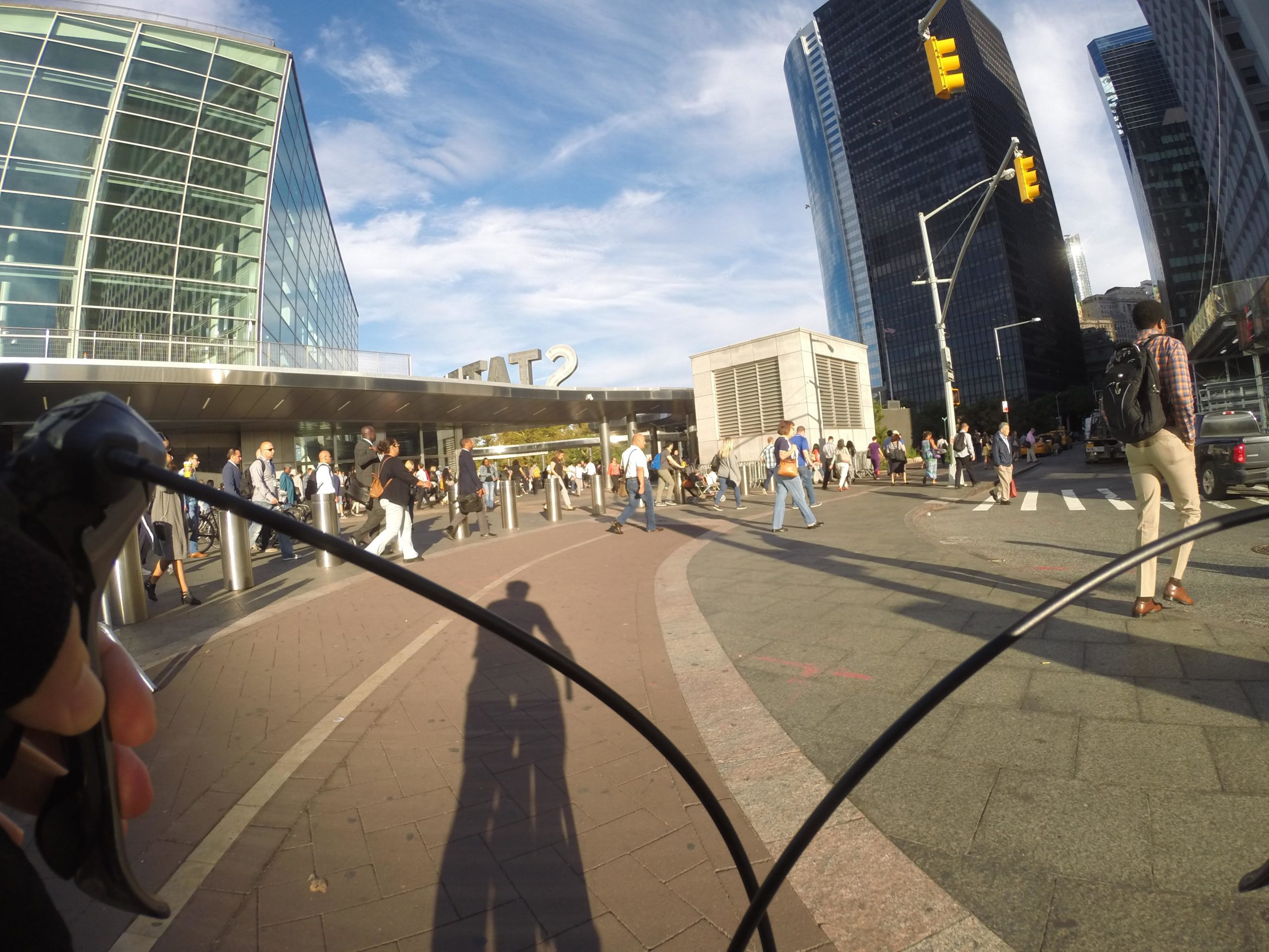 A bustling urban scene showing a group of people entering a modern glass-fronted building labeled "GATE 2." The image is taken from a cyclist's perspective, with bicycle handlebars partially in view. In the background, tall buildings reach towards the sky under a clear blue setting. Pedestrians are crossing the street, and there are vehicles on the road, adding to the lively city atmosphere. West Street Greenway mountain bike trail.