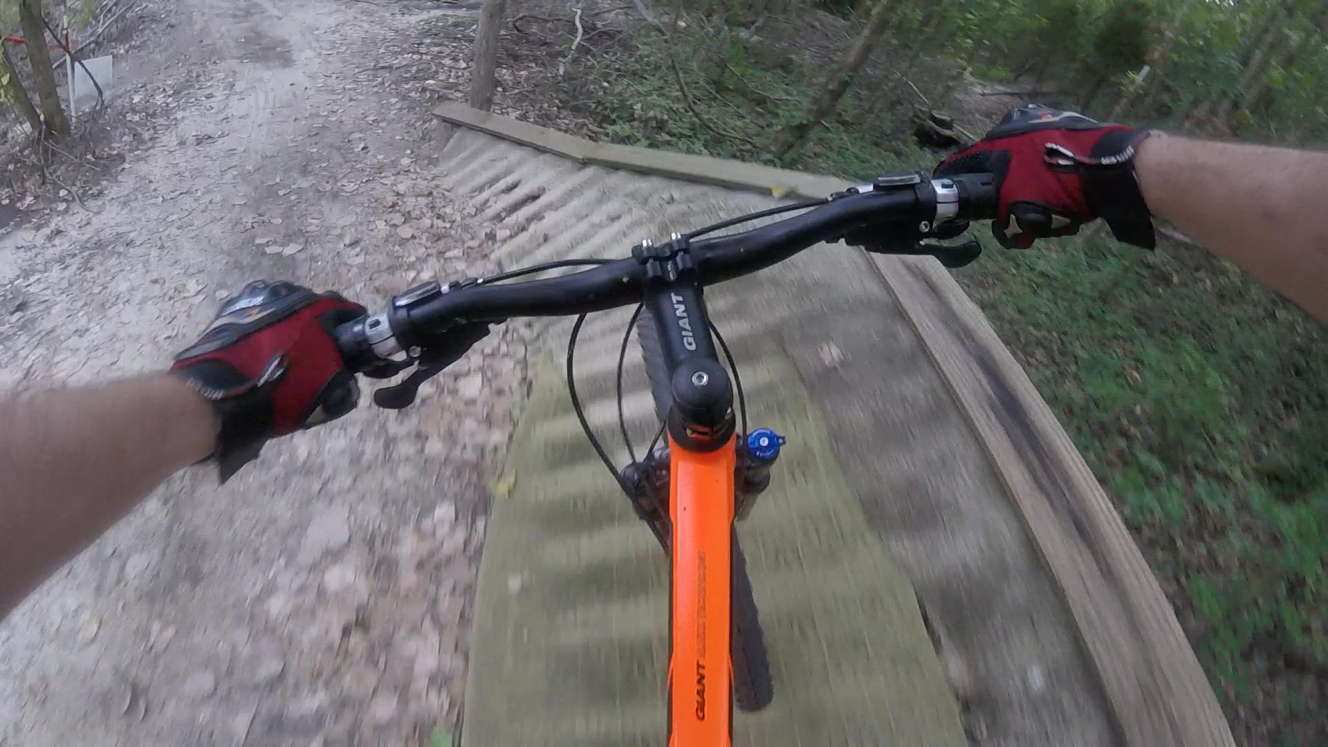 A close-up view of a mountain biker's hands gripping the handlebars of an orange bike while riding over a wooden ramp on a dirt trail surrounded by greenery. Horry County Bike Run Park mountain bike trail.