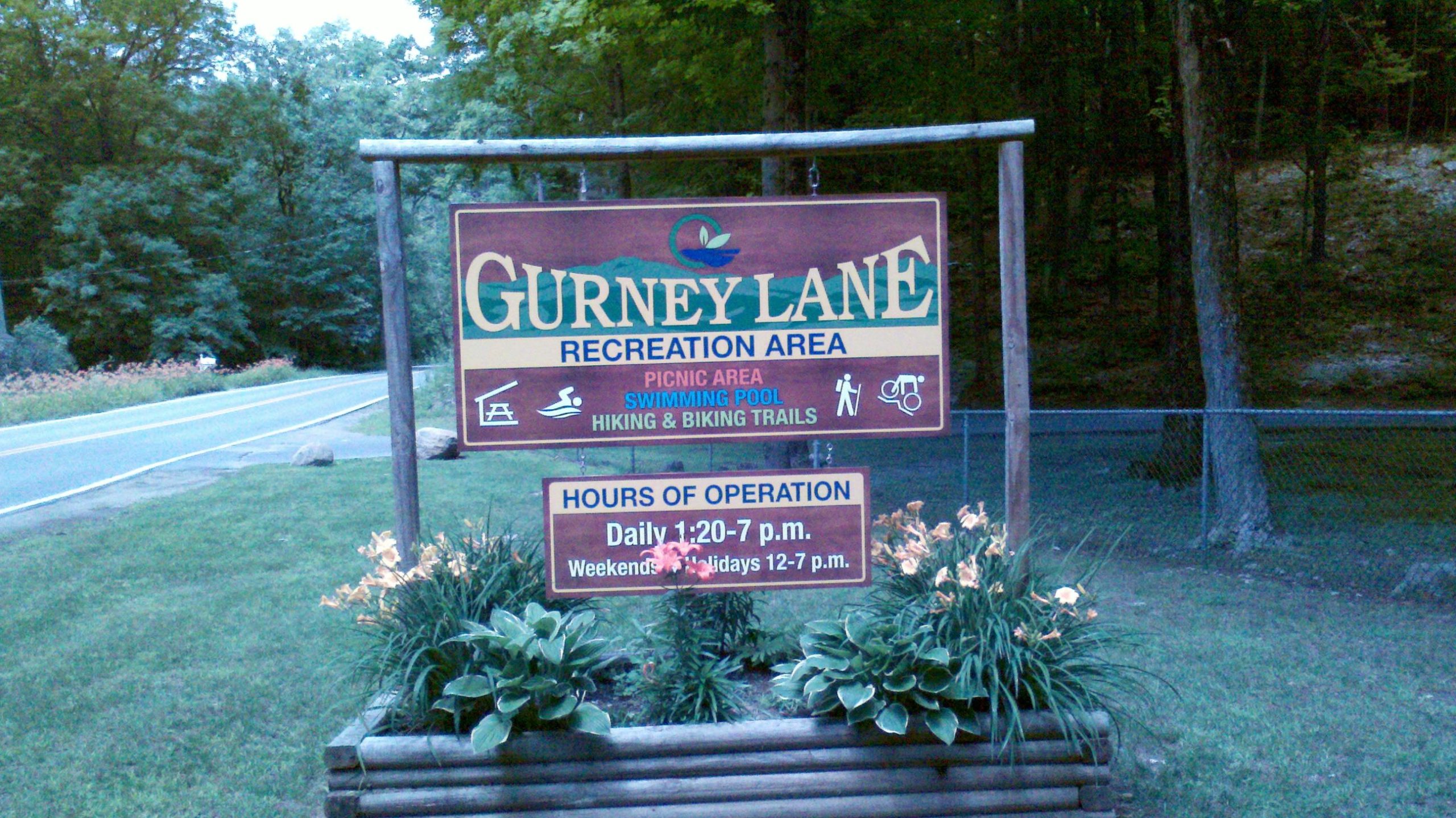 Image of a wooden sign for Gurney Lane Recreation Area, featuring information about the amenities available such as a picnic area, swimming pool, and hiking and biking trails. The sign includes hours of operation, indicating daily hours from 1:20 PM to 7 PM and weekends/holidays from 12 PM to 7 PM. The surrounding area is lush with greenery and flowers. Gurney Lane Mountain Bike Trails mountain bike trail.