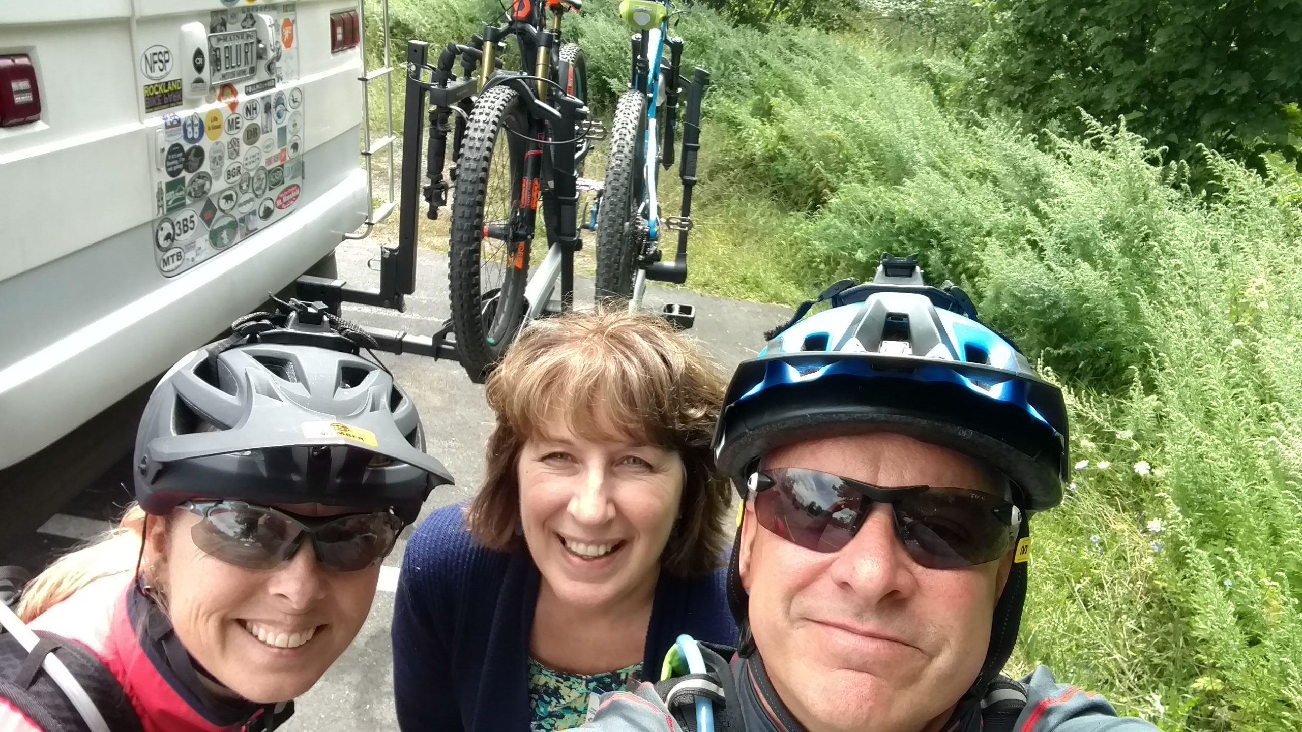 A group of three cyclists posing for a selfie in front of a camper van with numerous stickers. Two of them are wearing helmets and sunglasses, while the third person is smiling in a floral top. Mountain bikes are mounted on a bike rack behind them, surrounded by greenery. Pine Ridge mountain bike trail.