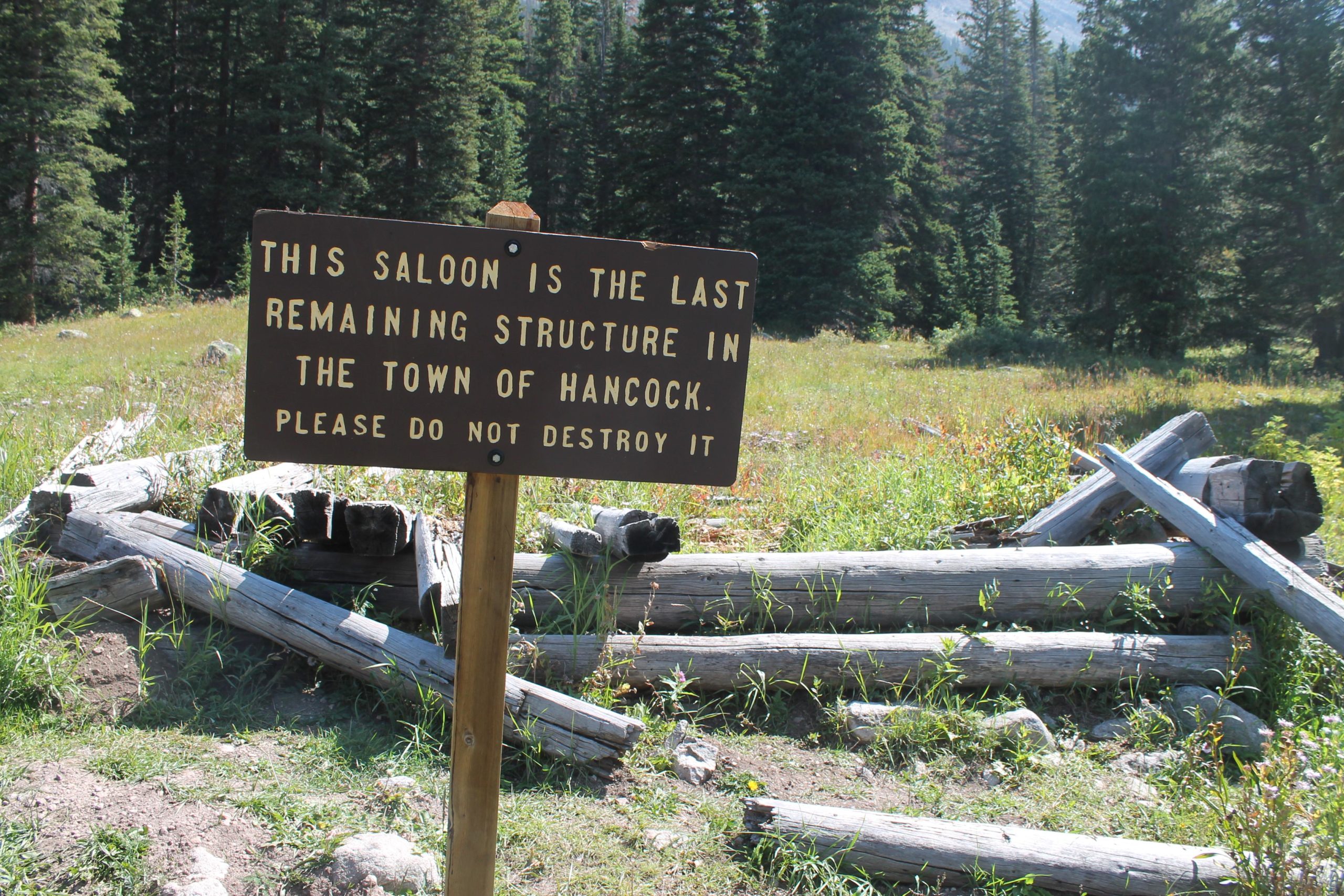 A wooden sign in a grassy area surrounded by trees, stating: "This saloon is the last remaining structure in the town of Hancock. Please do not destroy it." The area features some weathered logs and a natural landscape. Hancock Road mountain bike trail.