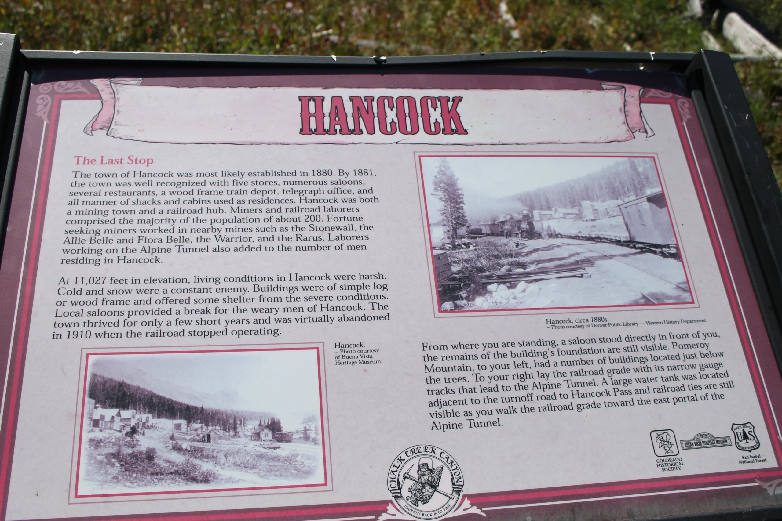 Information sign about Hancock, Colorado, detailing its history and development in the late 19th century. The sign includes text describing the establishment of Hancock in 1880, its population, living conditions, and economic activities related to mining and the railroad. It features historical photographs of the town and mentions notable buildings and features from the past. The sign is set against a natural backdrop, with grass and trees visible surrounding it. Hancock Road mountain bike trail.