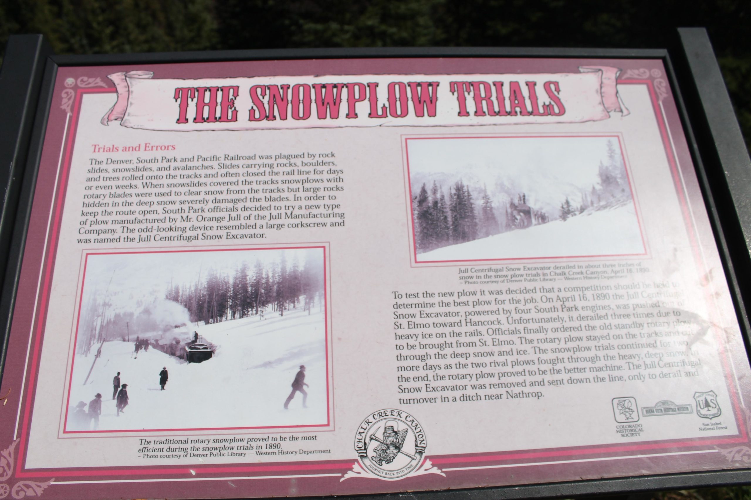 Sign describing the "Snowplow Trials," detailing the challenges faced by the Denver, South Park and Pacific Railroad due to snow and rock slides. The sign includes historical information about the trials and errors in snow removal methods, particularly highlighting the Jull Centrifugal Snow Excavator. It features a photograph illustrating early snowplow operation and includes a logo at the bottom. CDT: Tunnel Lake mountain bike trail.