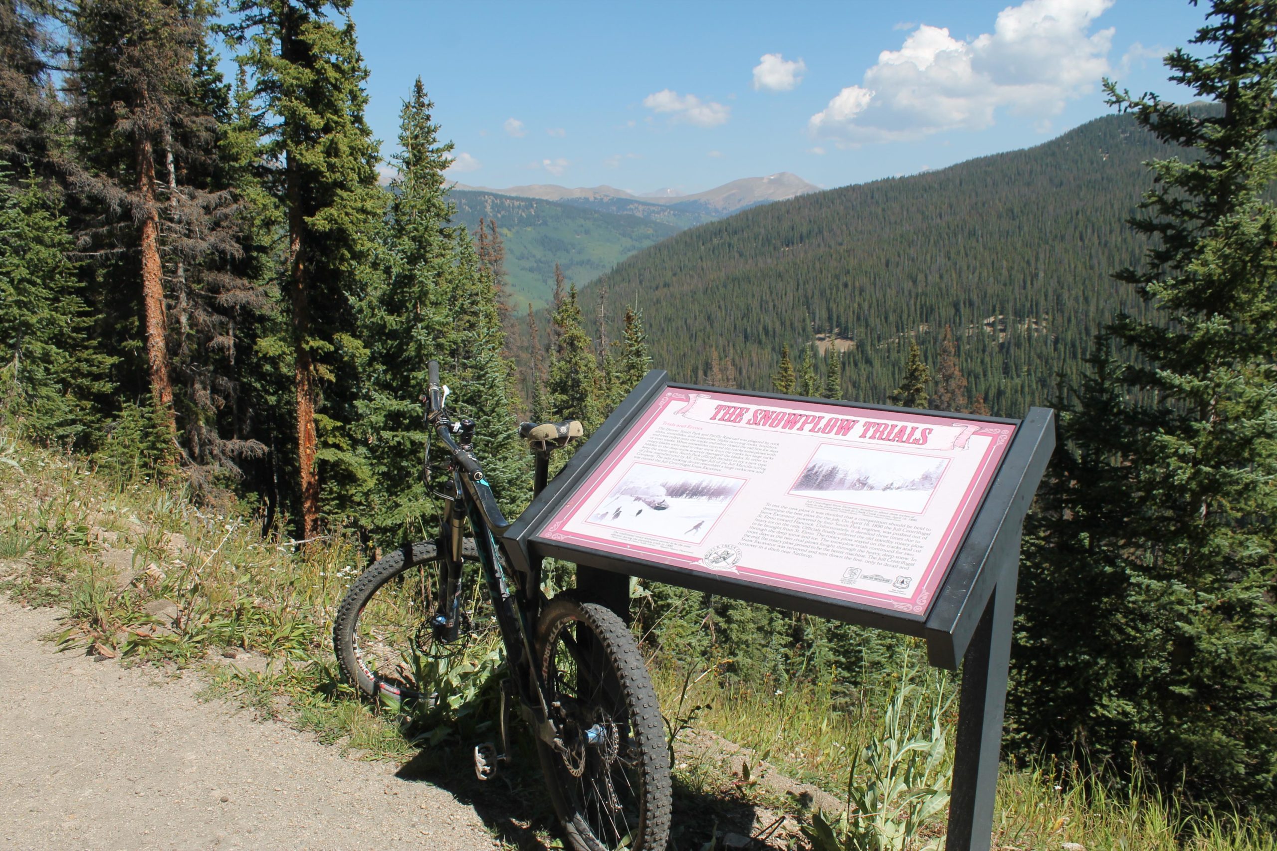A mountain bike parked beside an informational sign about the Snowplow Trails, surrounded by tall evergreen trees and a scenic mountainous landscape under a clear blue sky. CDT: Tunnel Lake mountain bike trail.