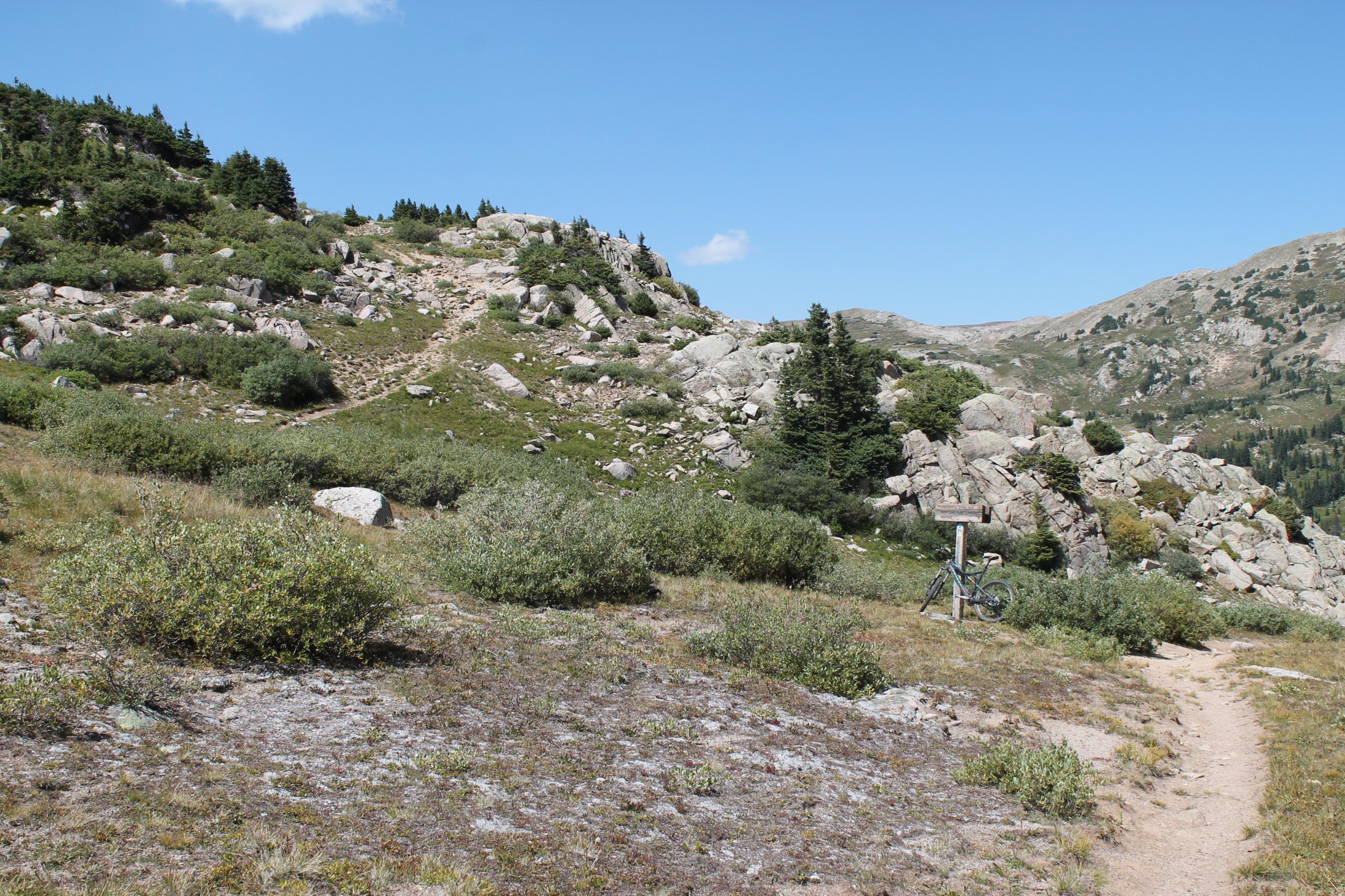 A rocky mountain trail surrounded by shrubs and low vegetation, with a grassy area in the foreground. A bicycle is leaning against a wooden trail sign, and the background features rolling hills and a blue sky with some clouds. CDT: Tunnel Lake mountain bike trail.