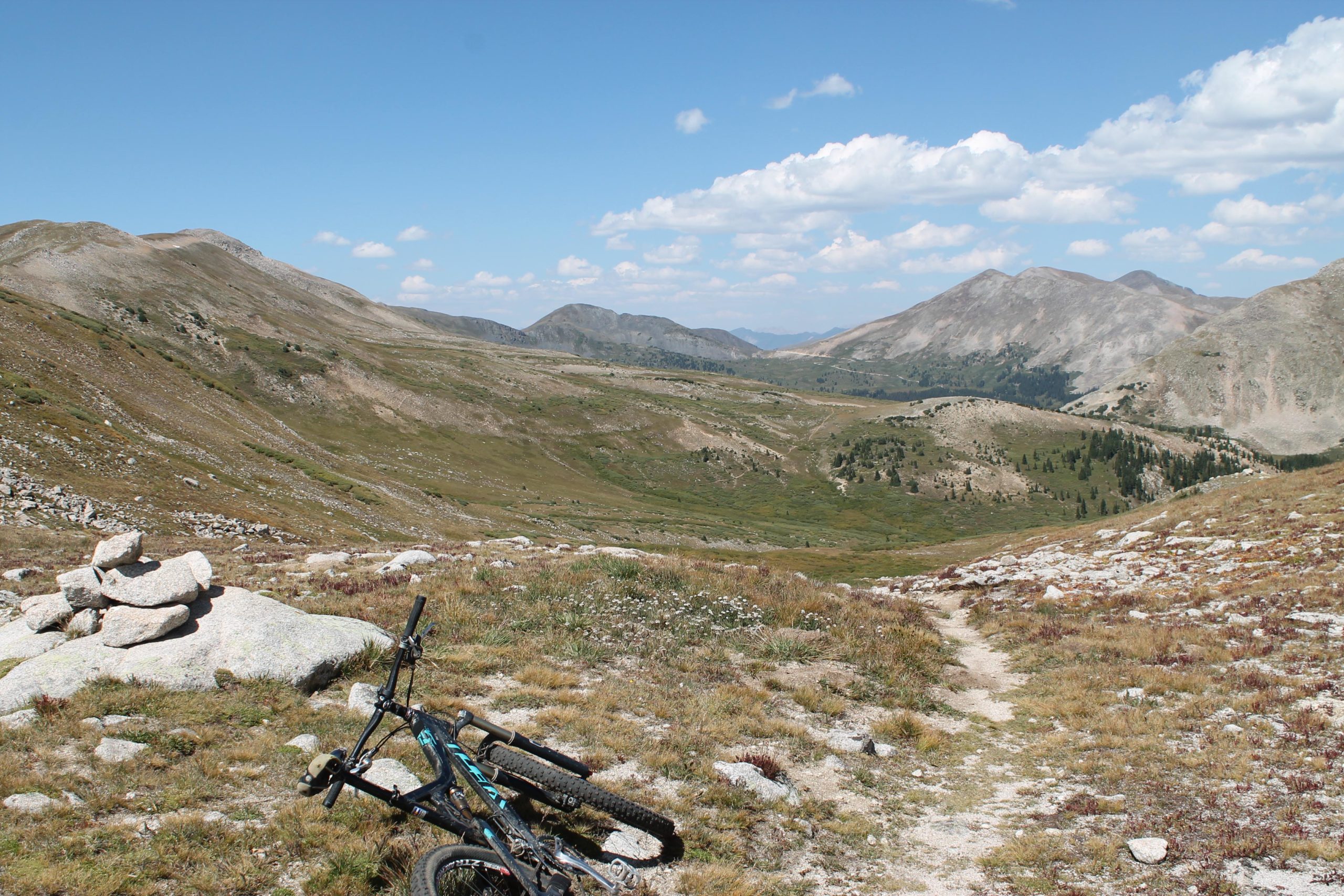 A mountain biking scene featuring a bike resting on rocky terrain, with a panoramic view of rolling hills and mountains under a blue sky with scattered clouds. A stone cairn is visible nearby, and the landscape is dotted with patches of grass and wildflowers. CDT: Tunnel Lake mountain bike trail.