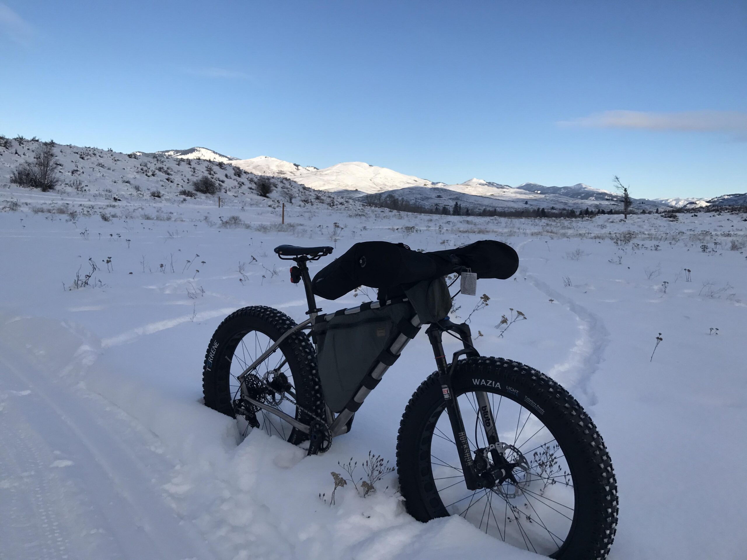 A fat tire bike rests on a snowy landscape, surrounded by a serene winter scene with distant mountains under a clear blue sky. The bike's tires are partially embedded in the snow, and tire tracks lead into the distance. Sparse vegetation peeks through the snow, enhancing the natural beauty of the wintry environment.