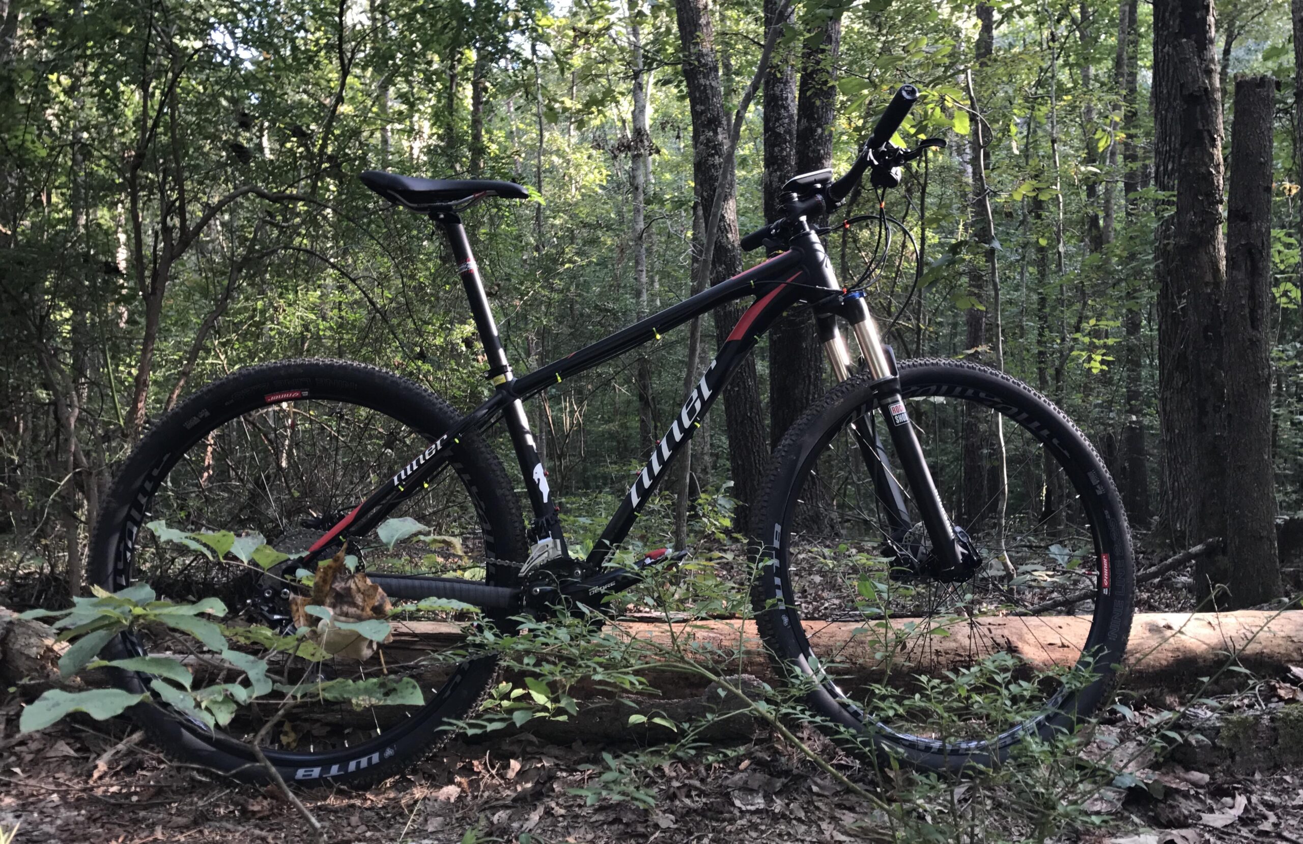 Niner EMD 9: A black mountain bike resting on a fallen log in a wooded area, surrounded by greenery and trees. The bike has thick tires and is positioned to showcase its frame and components, with sunlight filtering through the trees in the background.