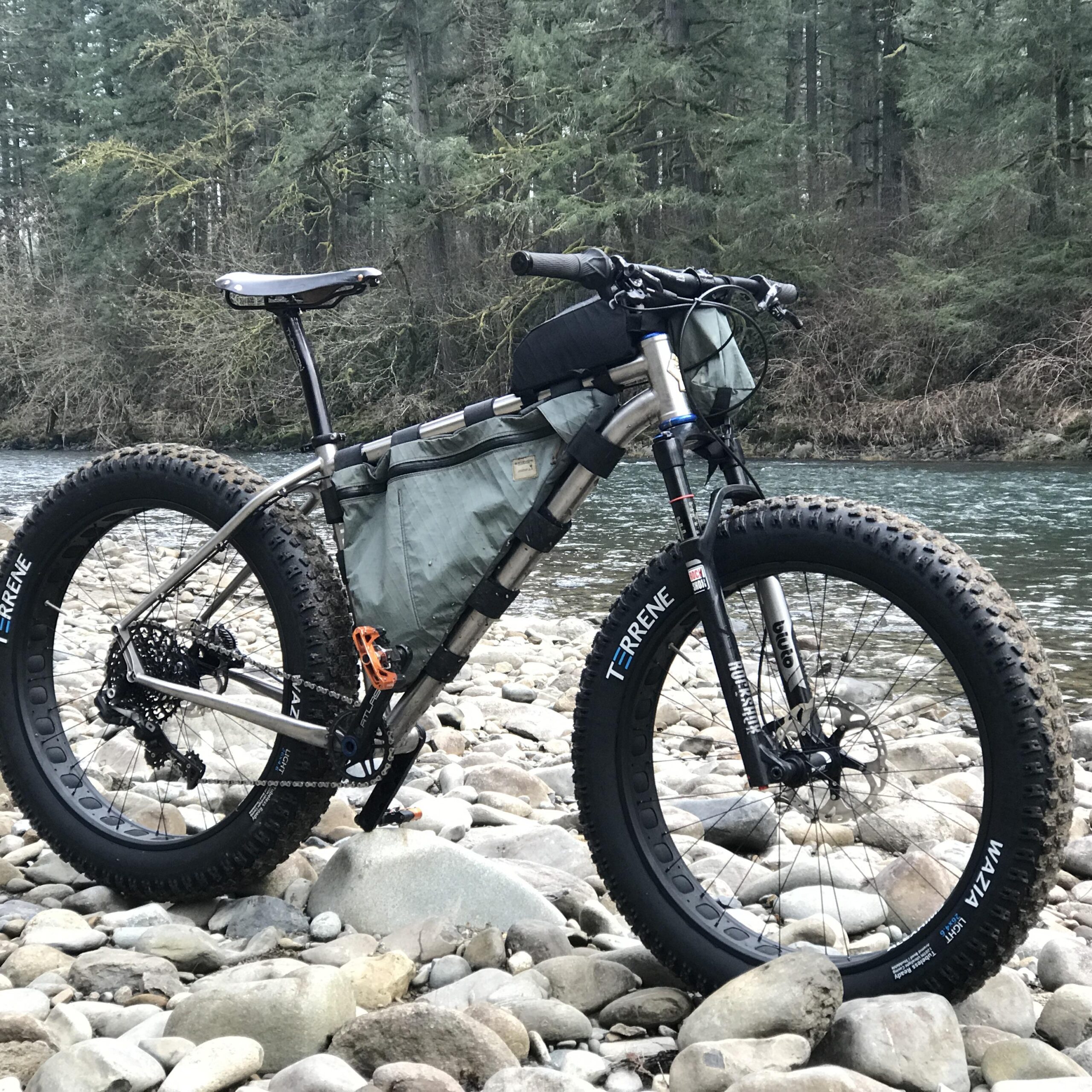 A rugged bike with fat tires and a gray frame rests on smooth stones by a riverbank, surrounded by lush green trees. The bike is equipped with a storage bag attached to the frame, suggesting it's designed for adventure or off-road cycling.