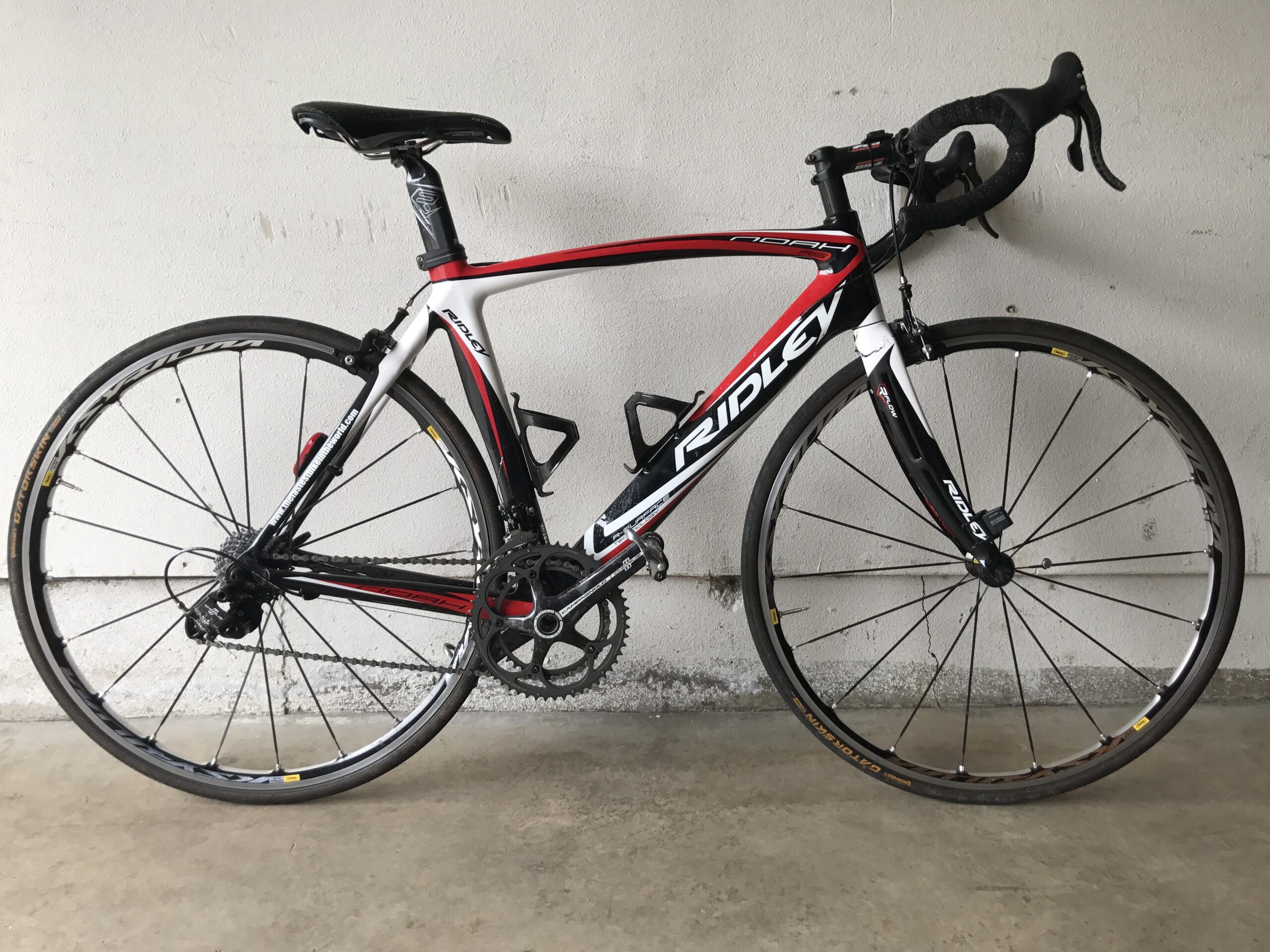 A red and white Ridley road bike with thin wheels and drop handlebars, positioned against a plain wall. The bike features a black saddle, gear components, and is displayed on a concrete surface.