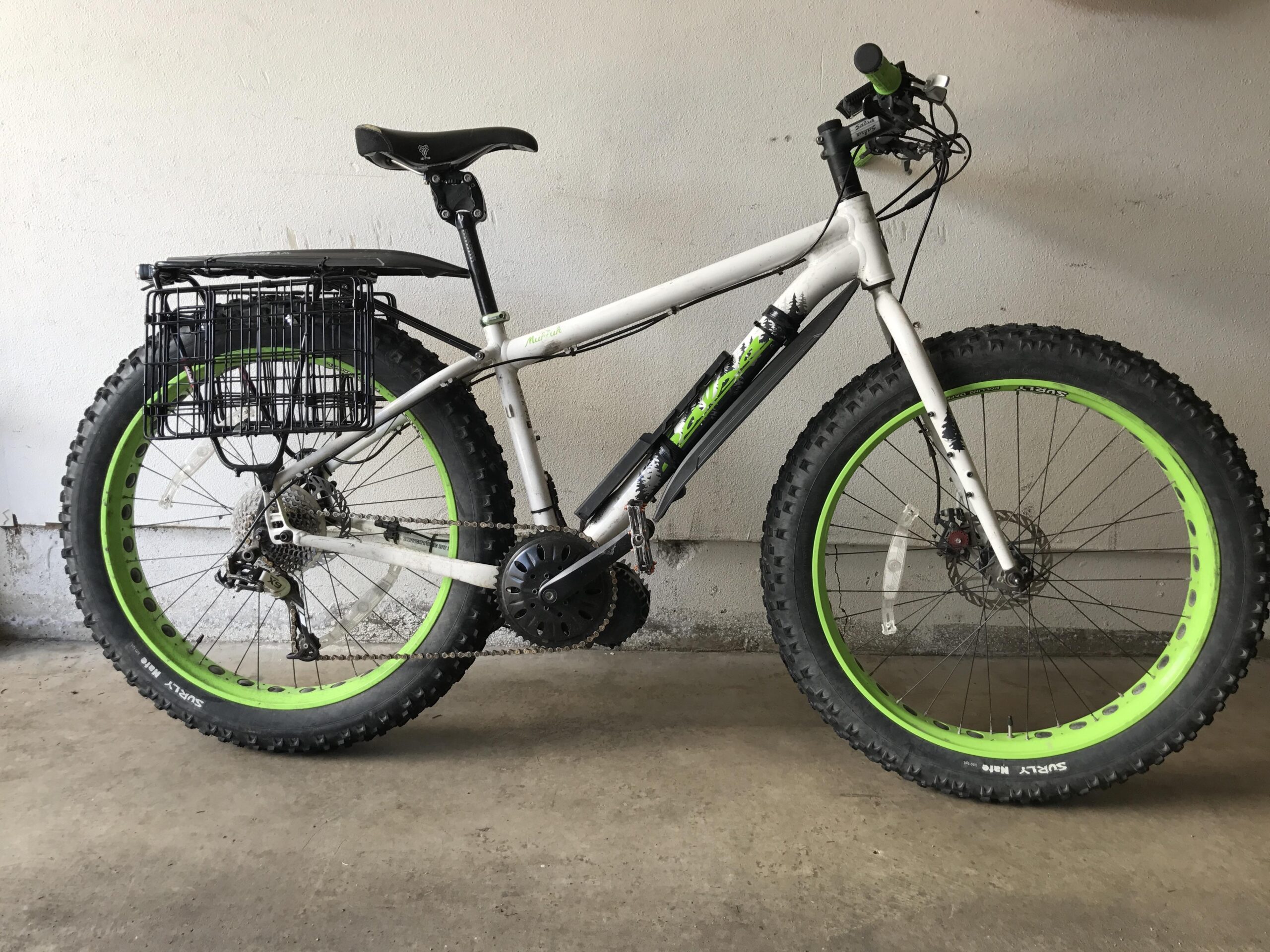 A white bicycle with green accents, featuring wide tires designed for off-road terrain. The bike has a black wire basket attached to the rear and a standard seat. It is positioned against a plain wall, highlighting the design and features of the bike.