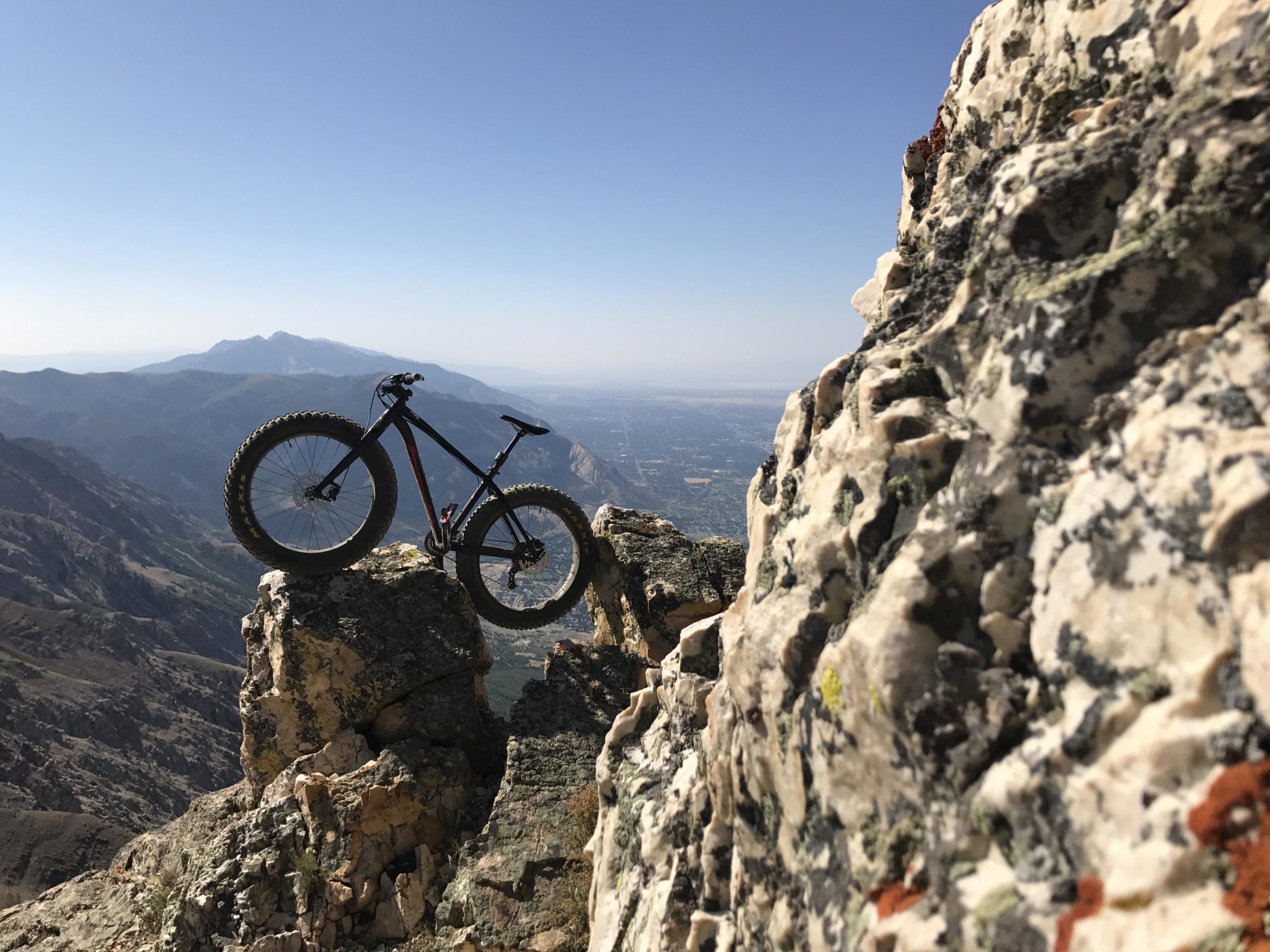 A mountain bike perched on a rocky outcrop, overlooking a scenic mountain landscape under a clear blue sky. The bike's large, rugged tires contrast with the rocky surface, while mountains fade into the distance. Northern Skyline mountain bike trail.