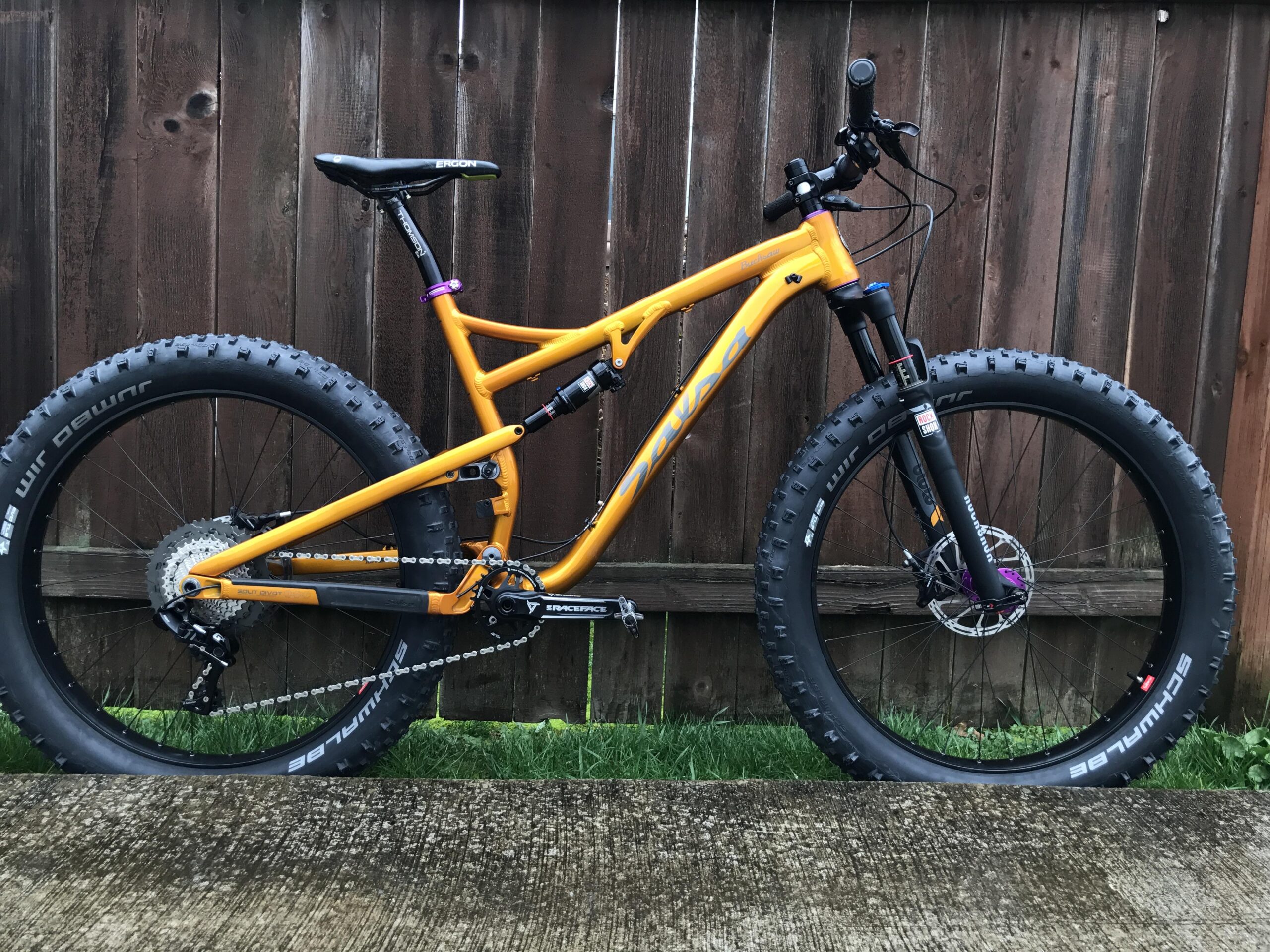 A bright orange mountain bike with fat tires positioned on a concrete surface. The bike features a modern frame design, adjustable seat, and front suspension. In the background, there is a wooden fence and some grass visible.
