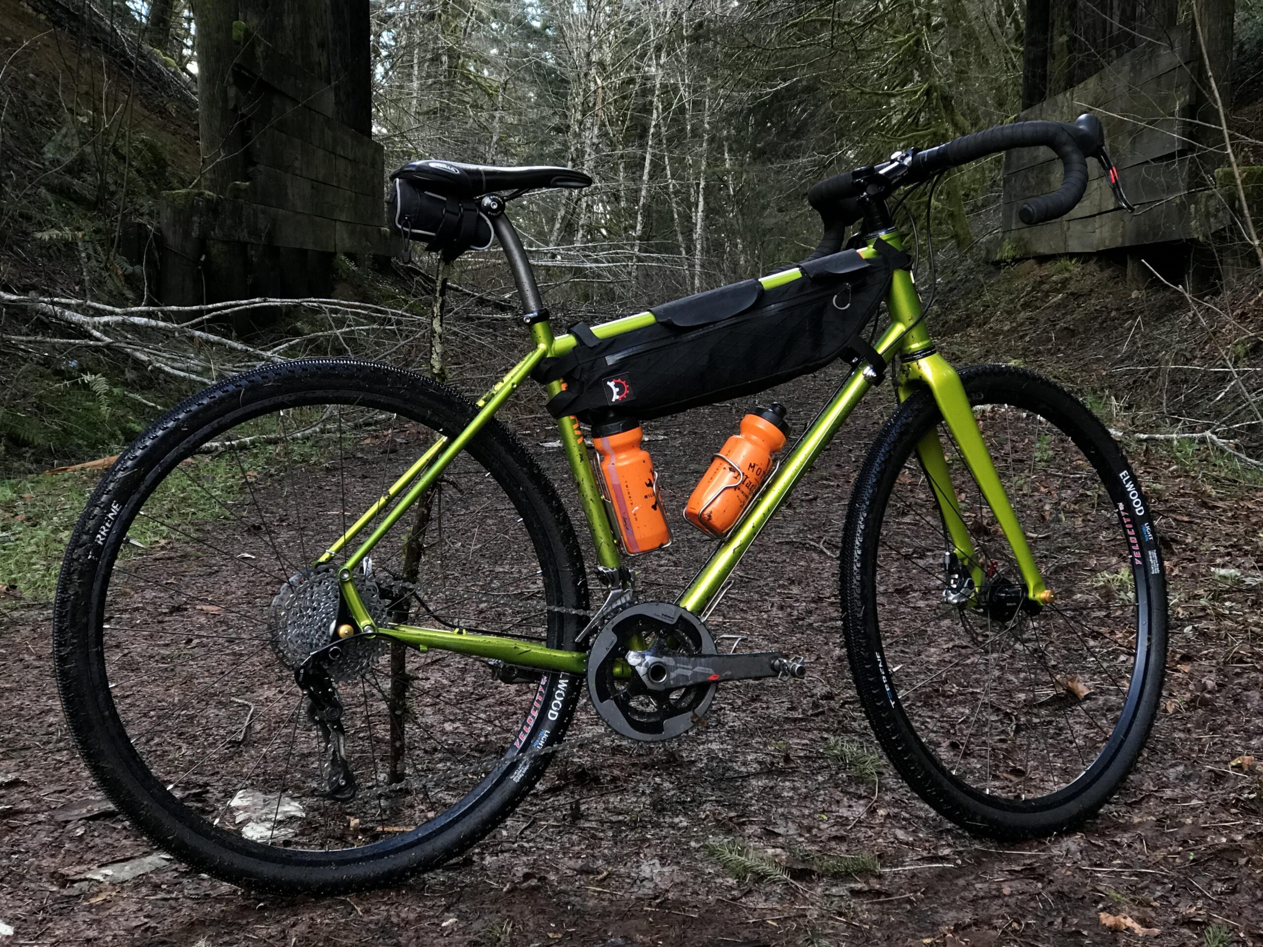 A mountain bike with a bright green frame is parked on a dirt trail surrounded by trees. It features a black bike bag mounted on the frame, and two bright orange water bottles secured in place. The background includes fallen branches and a rustic wooden structure, suggesting a natural, wooded area.