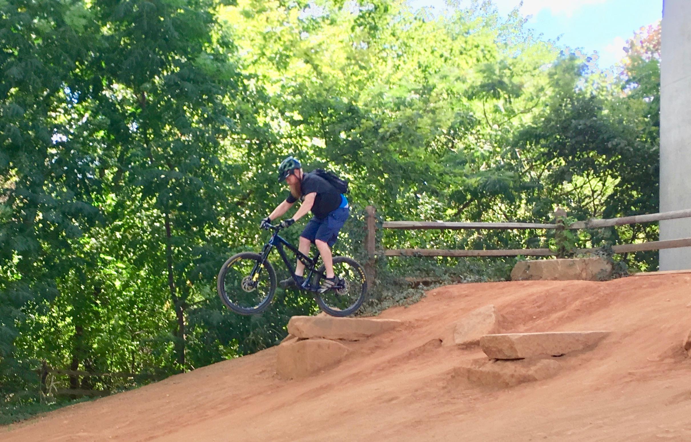 A person riding a mountain bike jumps off a small hill, surrounded by lush green trees and a wooden fence in the background. The mountain biker is wearing a helmet, a black shirt, and blue shorts, with a backpack on their back. Belle Isle mountain bike trail.