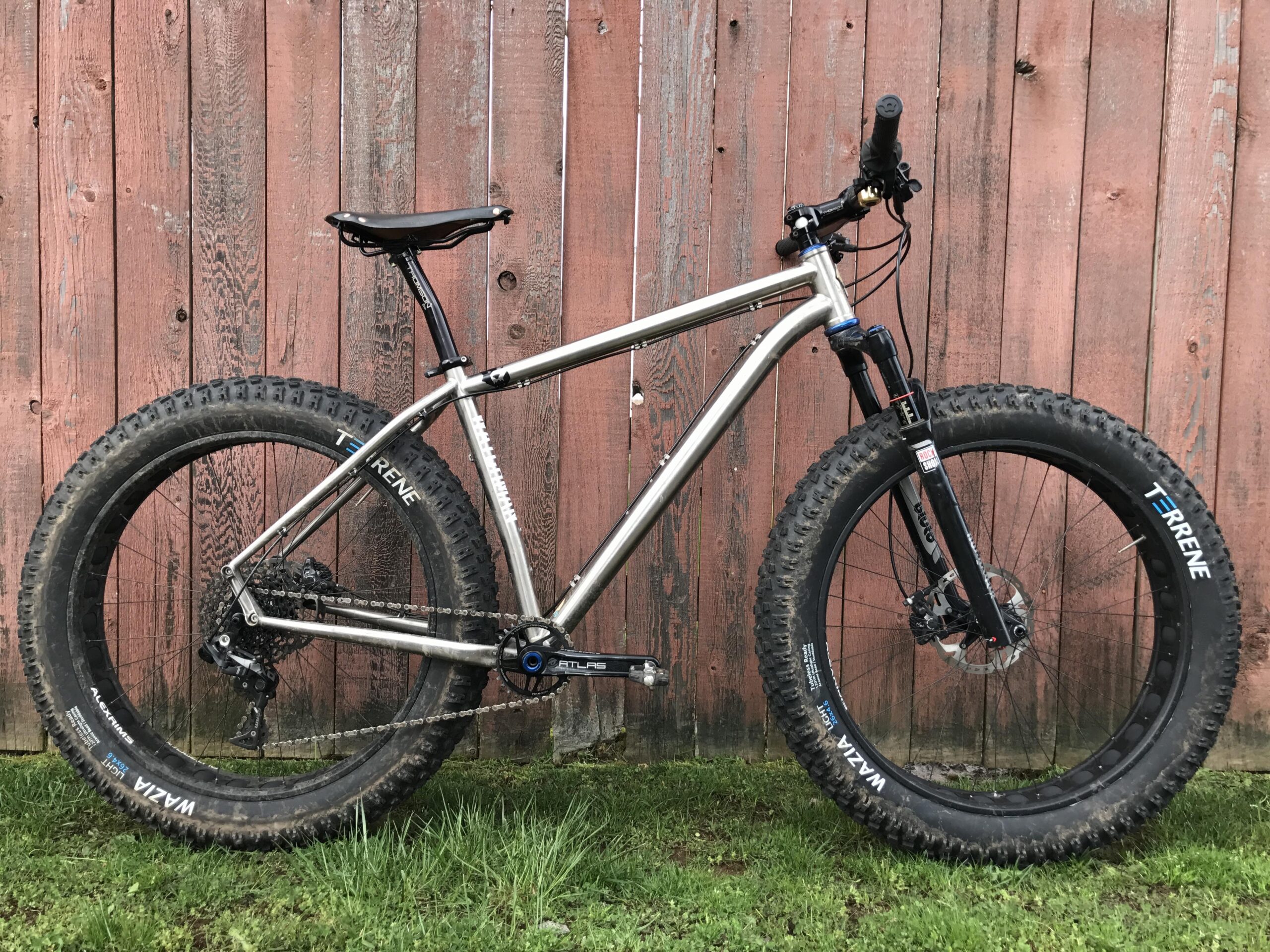 Alt text: A silver fat bike standing on grass in front of a rustic wooden fence. The bike features wide tires suitable for off-road riding, a black suspension fork, and a sleek design with a minimalist frame and seat.