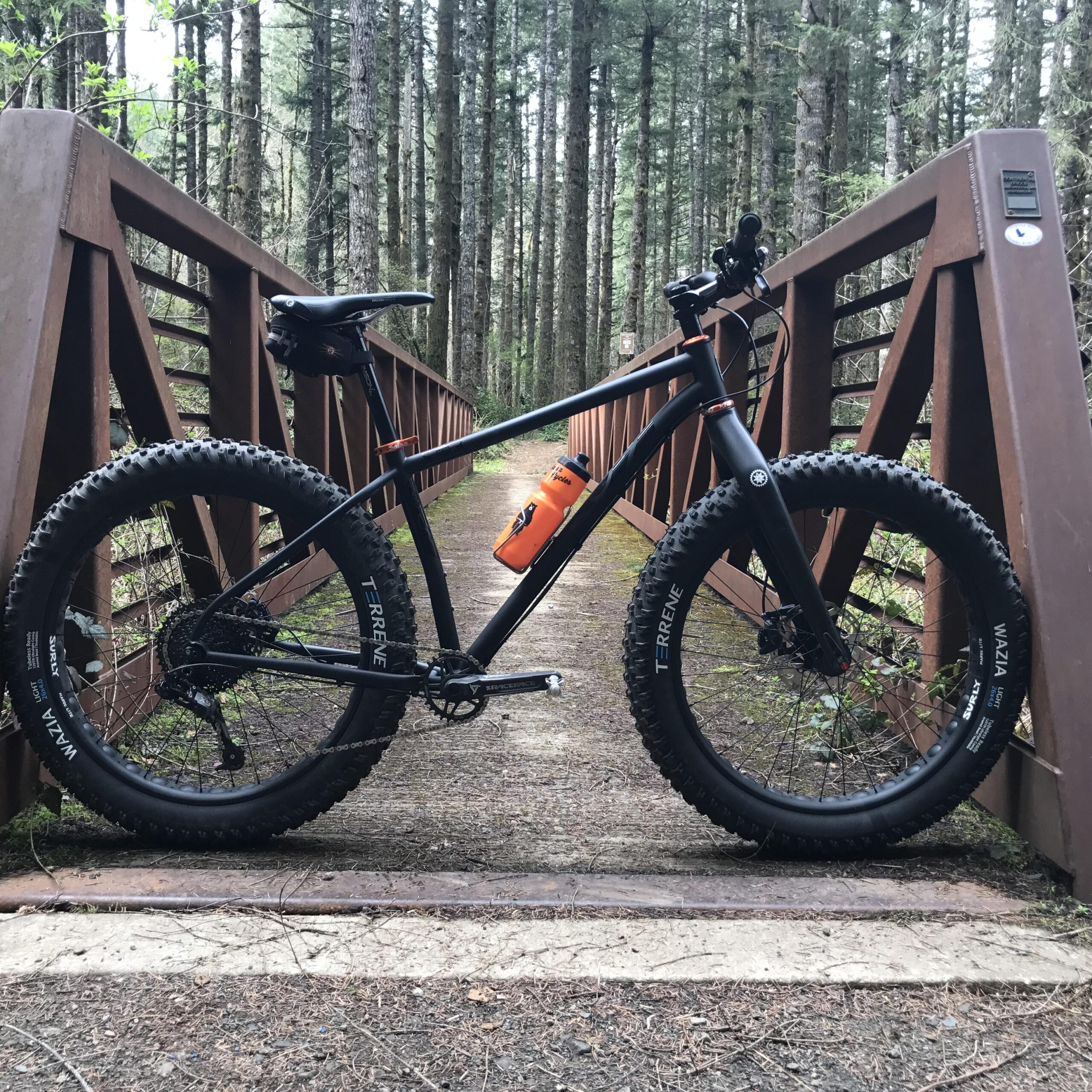 A black fat bike with orange accents parked on a wooden bridge surrounded by tall trees in a forest setting. The bike features wide tires and has an orange water bottle attached to its frame.