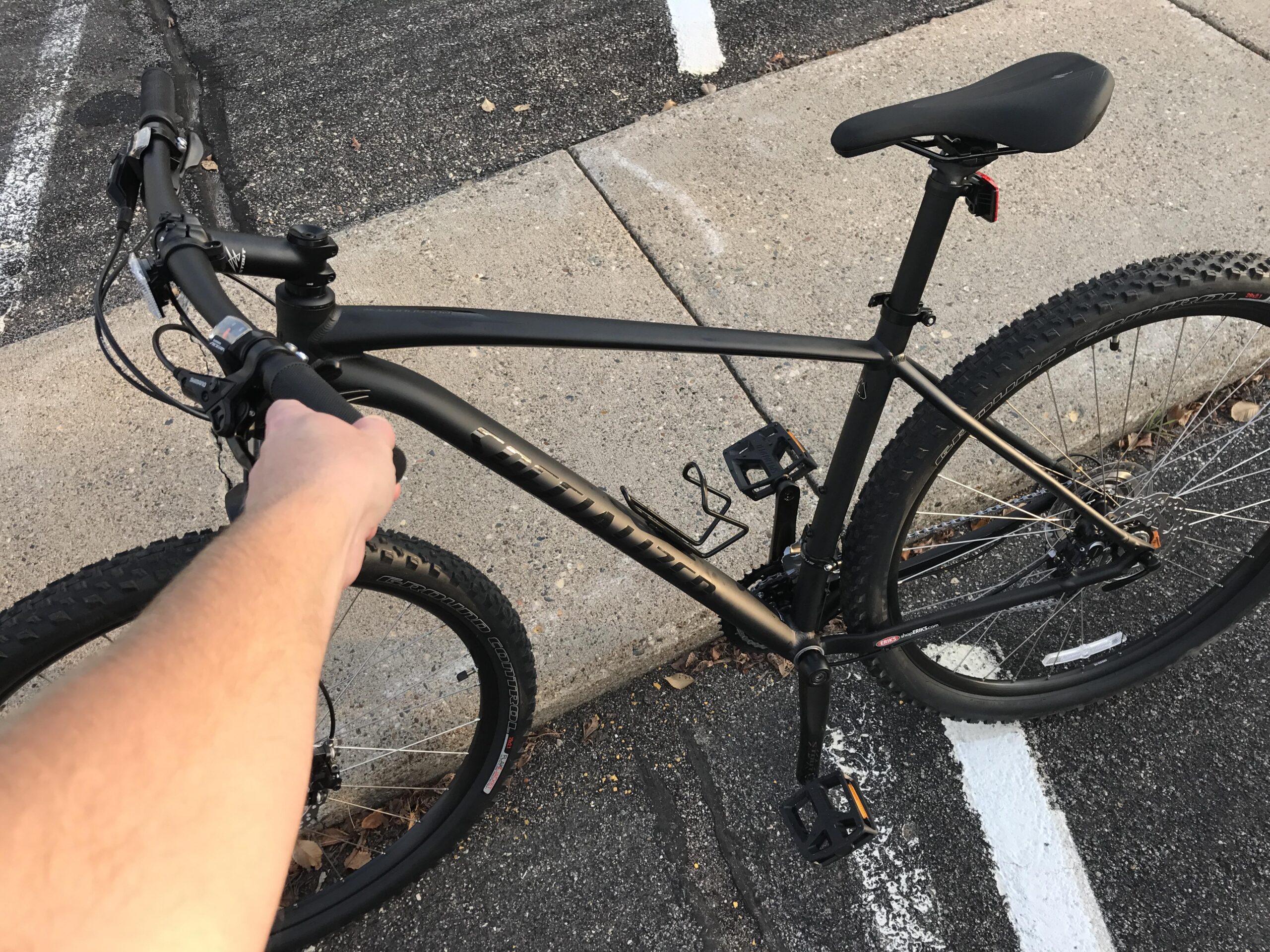 Specialized Rockhopper Comp 29: A close-up view of a mountain bike held by a person's hand. The bike features a matte black frame, black handlebars, and knobby tires. The background shows a partially visible concrete curb and a striped parking area.