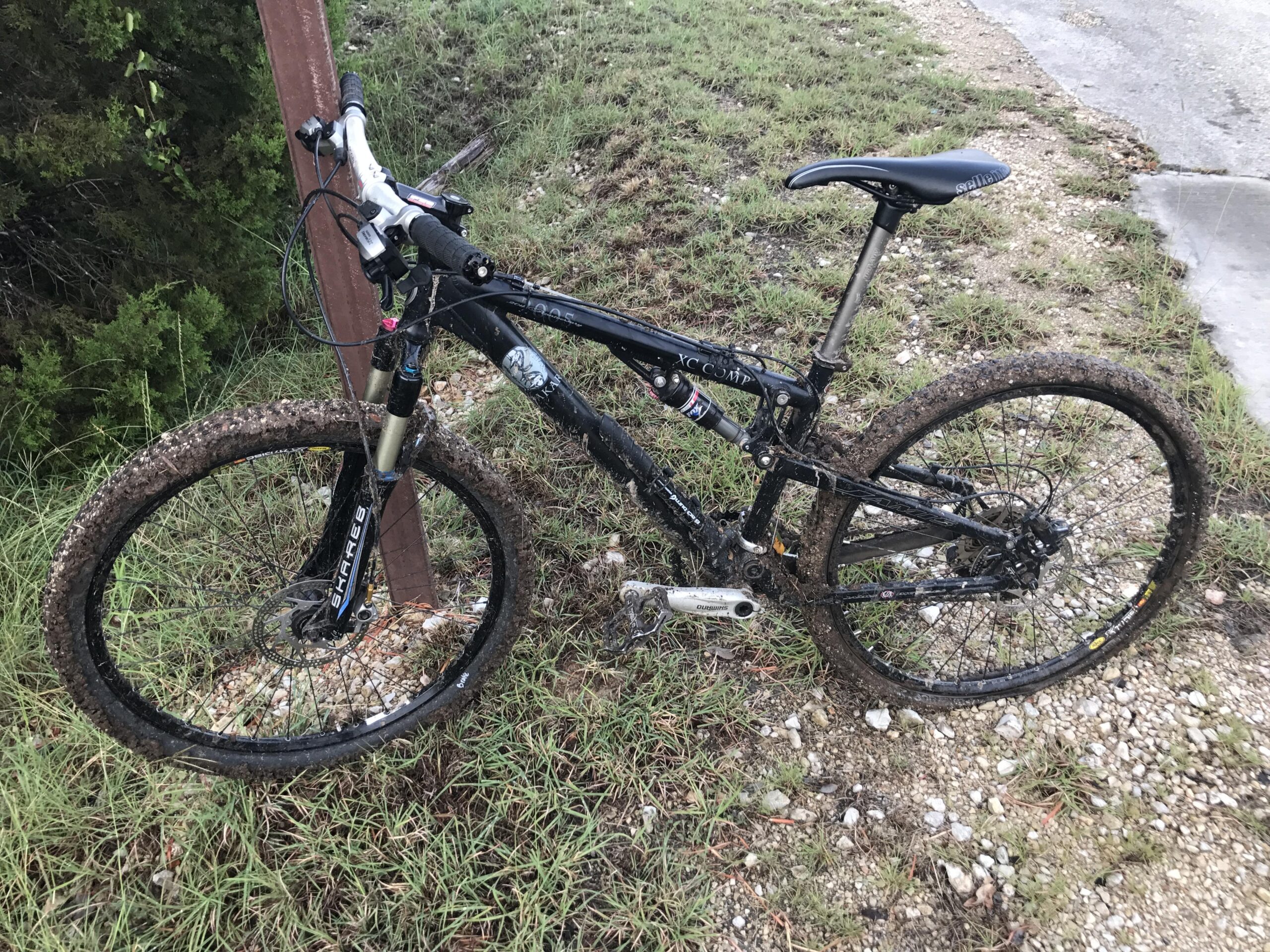 Jamis Dakar XC: A black mountain bike with muddy tires is parked next to a rusty metal pole on a grassy area. The background features some greenery, and there is gravel and a paved path nearby.