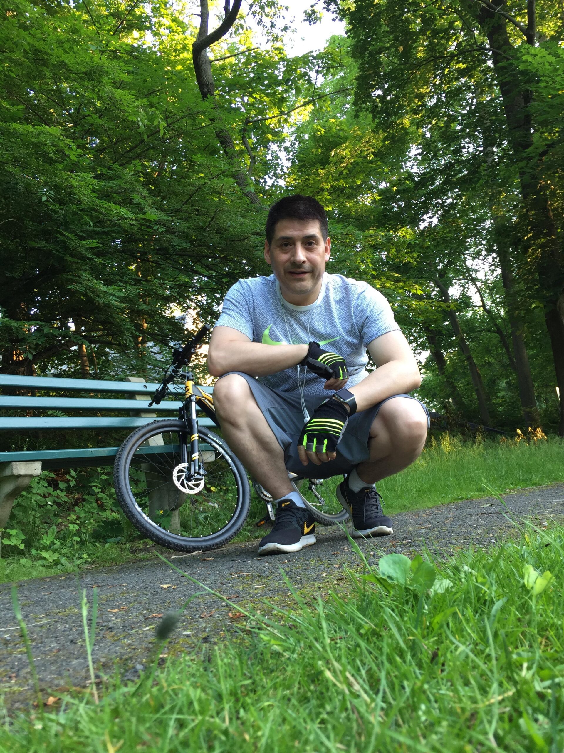 2017 Giant Talon 3: A man crouches next to a mountain bike on a scenic path, surrounded by lush green trees. He is wearing a light gray athletic shirt, shorts, and black sneakers with yellow accents. The setting is bright and natural, showcasing a peaceful outdoor environment.