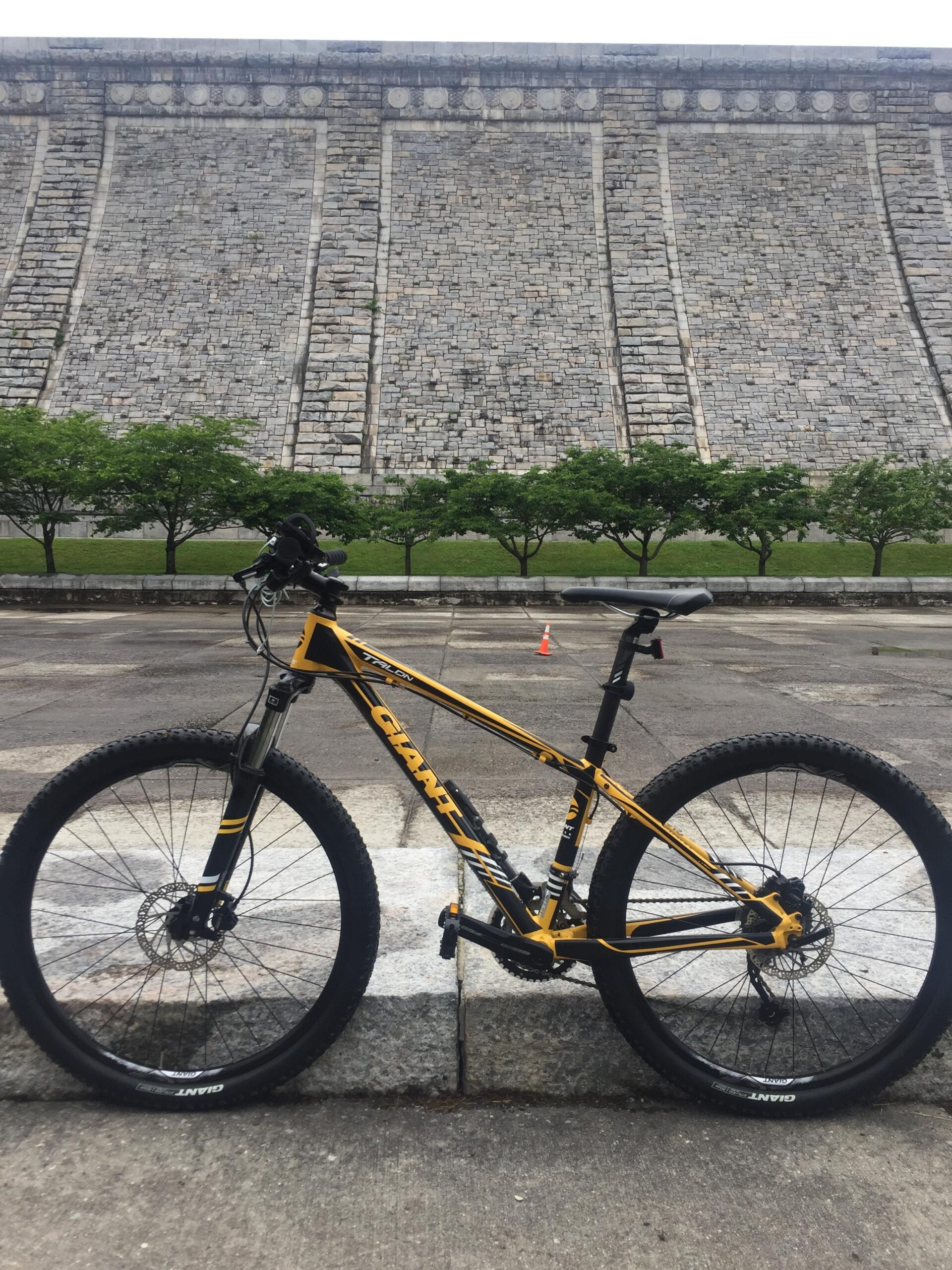 2017 Giant Talon 3: A yellow and black mountain bike is positioned in front of a large stone dam. The bike features thick tires and a sturdy frame, resting on a paved area. In the background, the dam's textured stone wall rises steeply, and small green trees line the base of the dam. A small orange cone is visible on the ground in the distance.