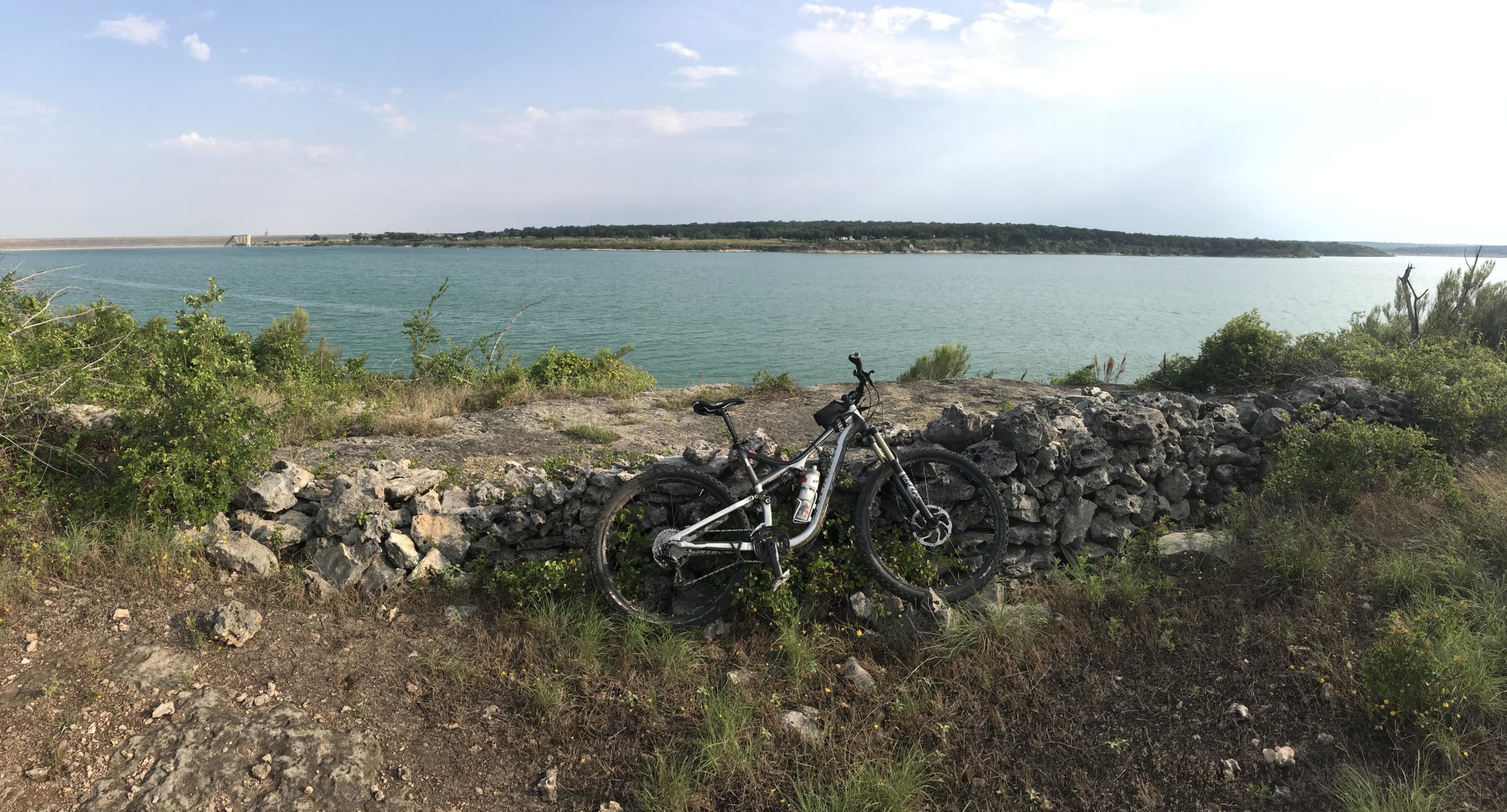A mountain bike leaning against a stone wall beside a tranquil body of water, surrounded by greenery under a partly cloudy sky. Goodwater Trail mountain bike trail.
