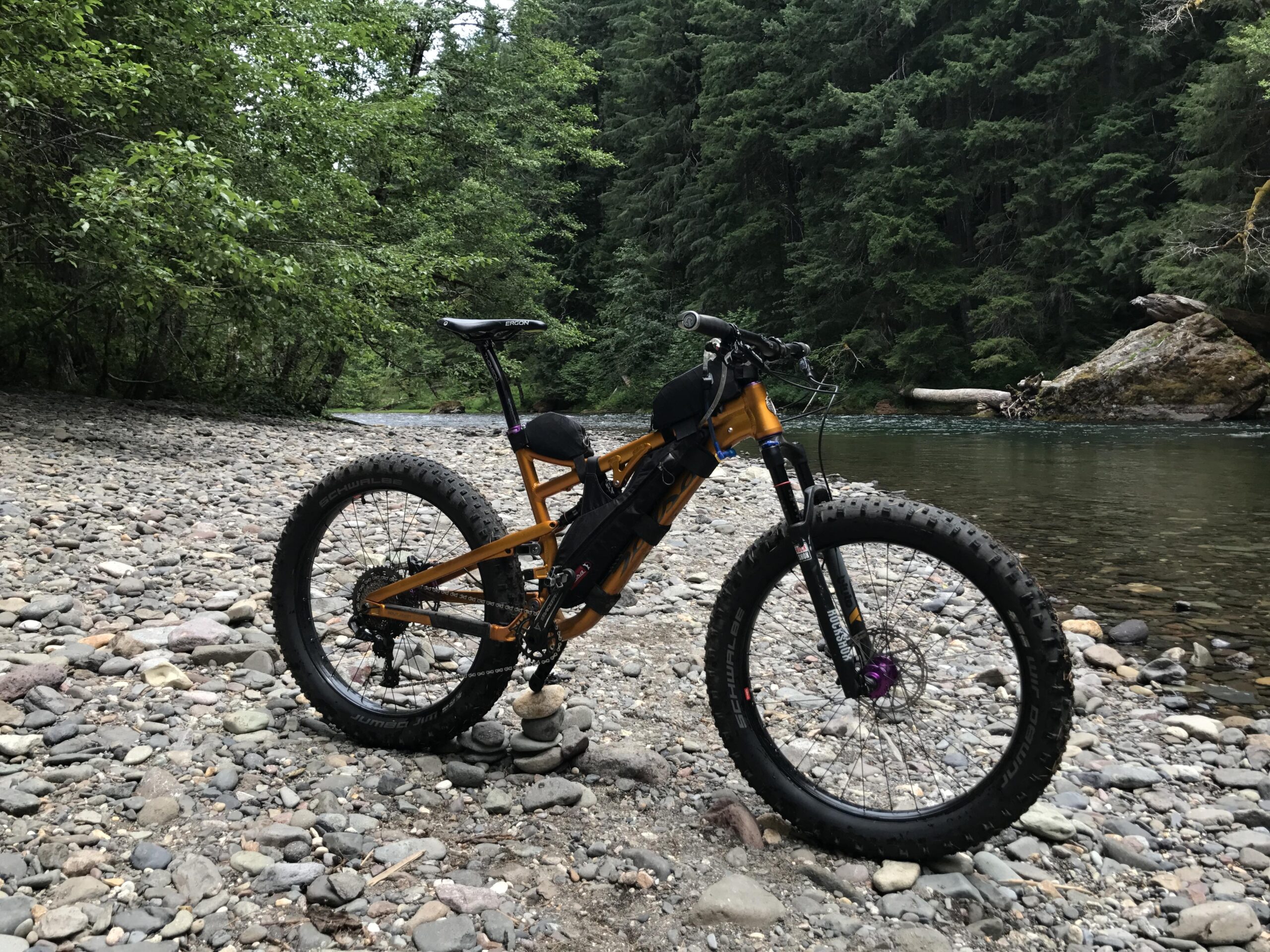A fat tire bike parked on a rocky riverbank, surrounded by lush green trees and a flowing stream in the background. The bike features an orange frame, large tires, and a black storage pack attached to the frame.