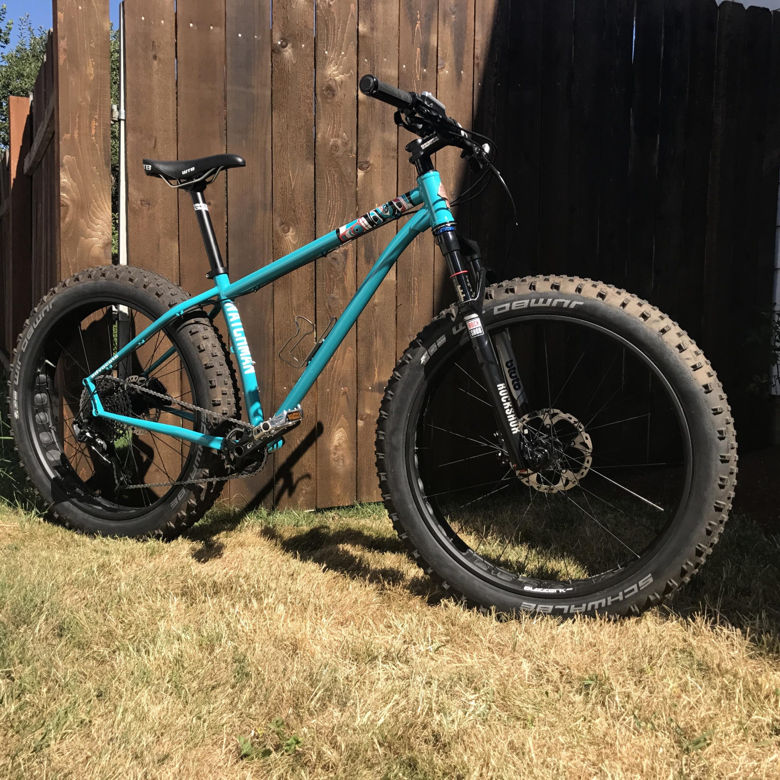 A teal mountain bike with oversized tires stands on dry grass, leaning against a wooden fence. The bike features a sturdy frame, black handlebars, and a noticeable suspension fork, showcasing its design for rugged terrain.