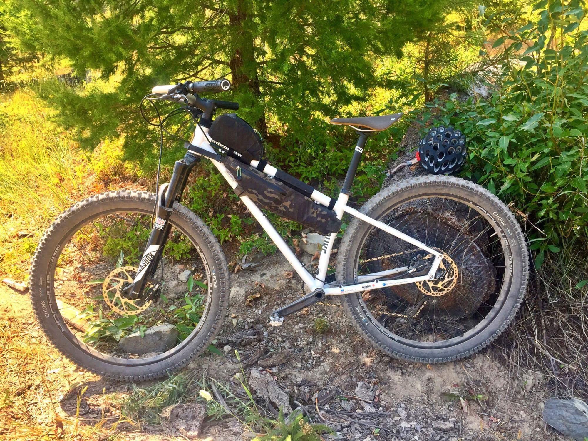 A mountain bike positioned on a gravel path surrounded by greenery. The bike features a black and white frame, thick tires, and a small bag attached to the frame. A black helmet is placed next to the bike, with sunlit foliage in the background.