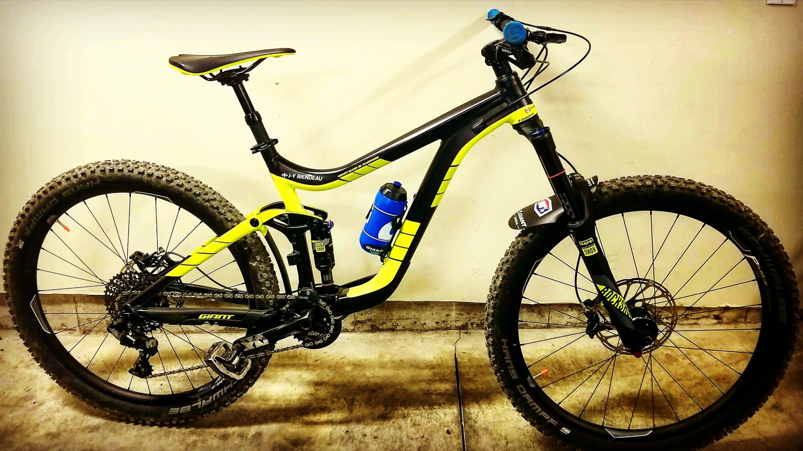 Giant Reign 2: A black and yellow mountain bike leaning against a light-colored wall. The bike features large, knobby tires, a durable frame, and a water bottle holder. The bike's components include a suspension fork, disc brakes, and a geared drivetrain, showcasing its suitability for rugged trails.