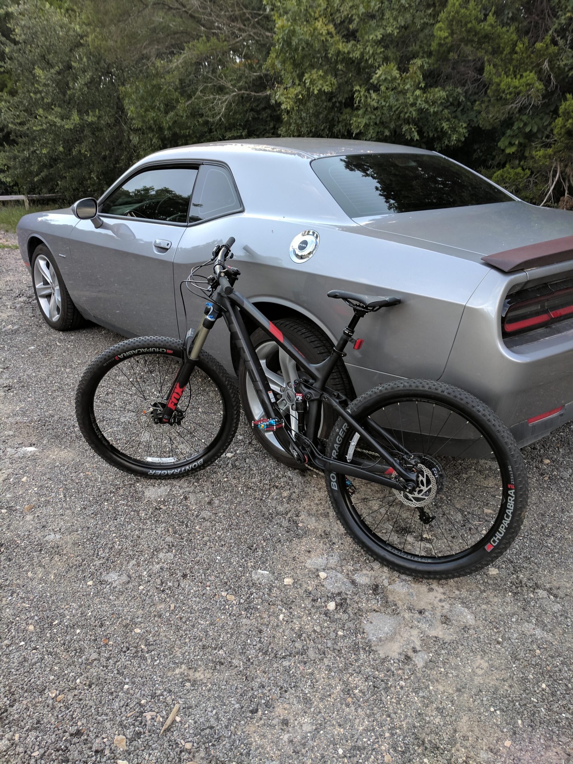 Trek Fuel EX 8: A black mountain bike stands next to a silver sports car parked on gravel, surrounded by lush greenery. The bike is equipped with thick tires and front suspension, while the car features a sleek design with distinctive rear lights.