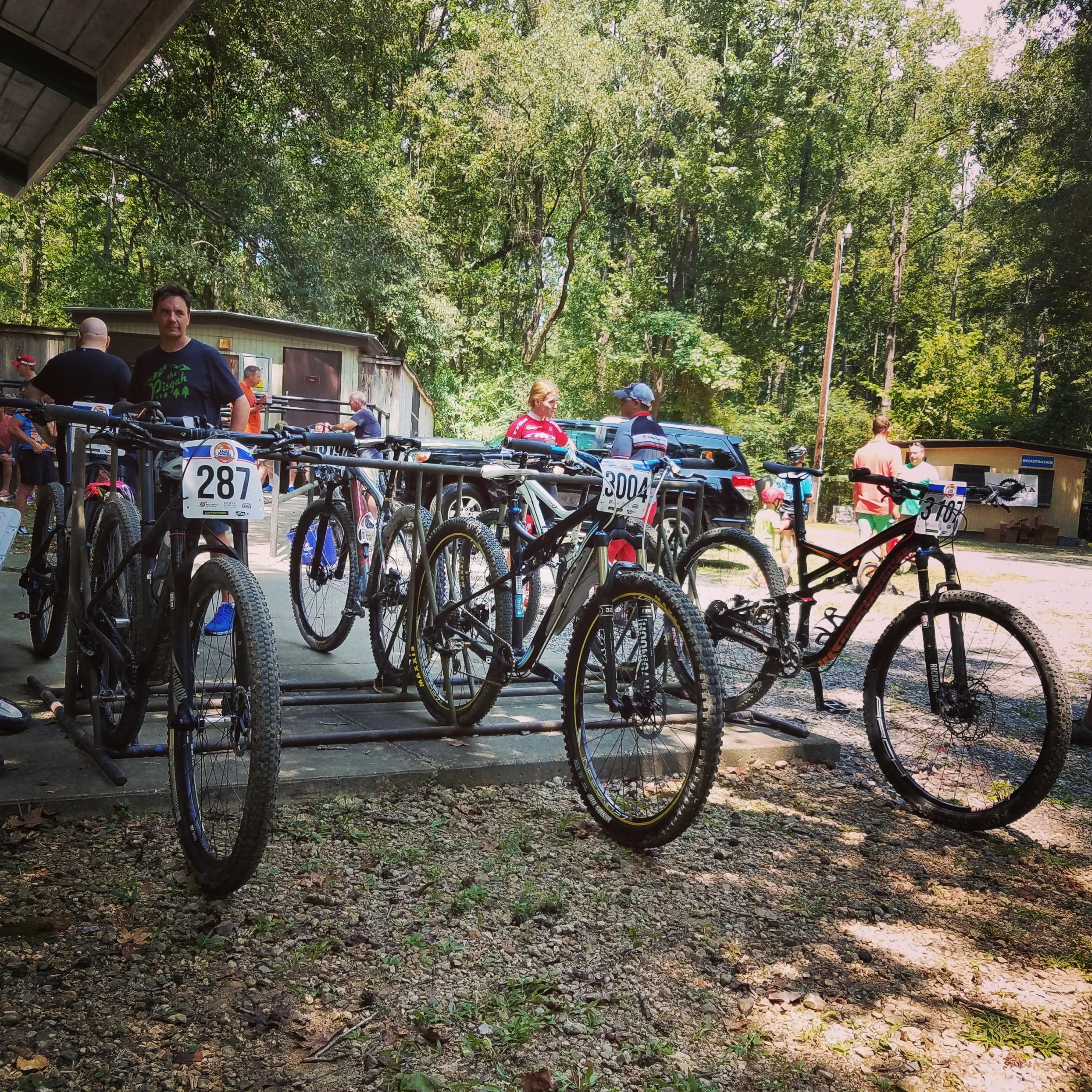 A group of mountain bikes secured on a bike rack, with several riders and spectators chatting in the background. The setting is outdoors, surrounded by trees, and shows a mix of enthusiasm and camaraderie among the participants at a biking event. Some bikes have race numbers attached. Comite Trails mountain bike trail.