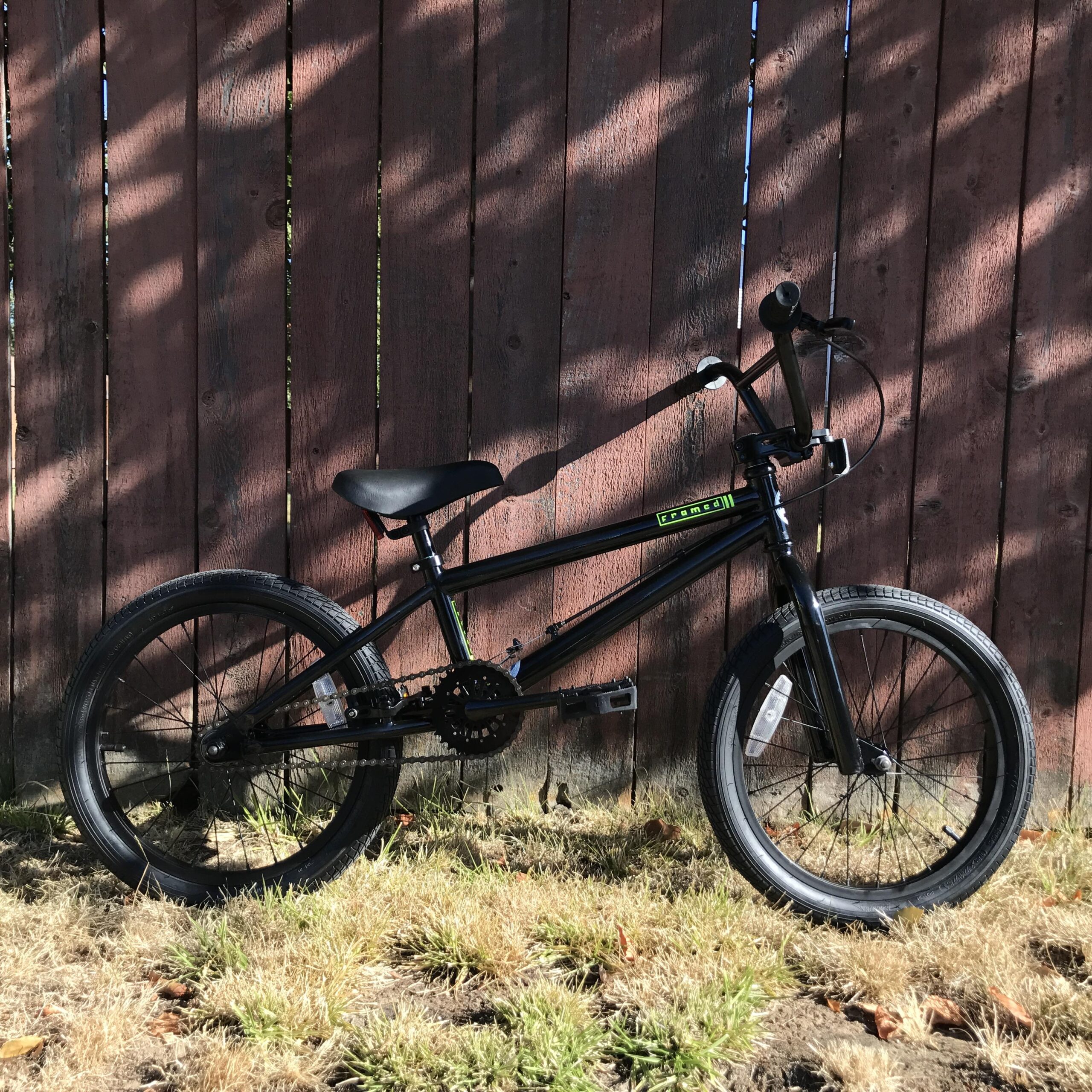 A black BMX bike stands on a patch of grass in front of a weathered wooden fence. The bike features a sleek frame, a comfortable seat, and sturdy tires, showcasing a design suited for tricks and stunts. Sunlight casts shadows on the fence, creating an outdoor atmosphere.