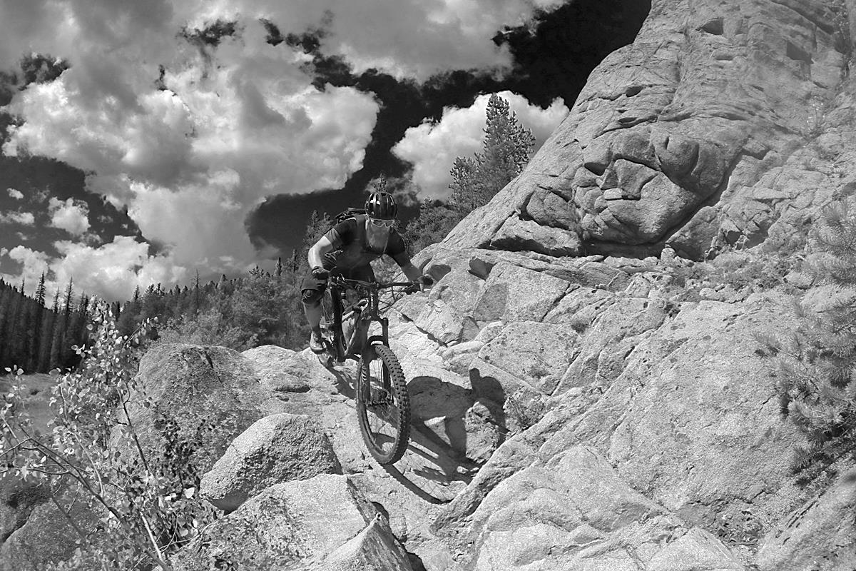 A mountain biker navigating rocky terrain under a cloudy sky, showcasing a dynamic outdoor adventure. The scene is captured in black and white, highlighting the rugged landscape and the cyclist's focused effort as they maneuver over large stones and boulders. Pine trees are visible in the background, adding to the natural setting. Canyon Creek Trail mountain bike trail.