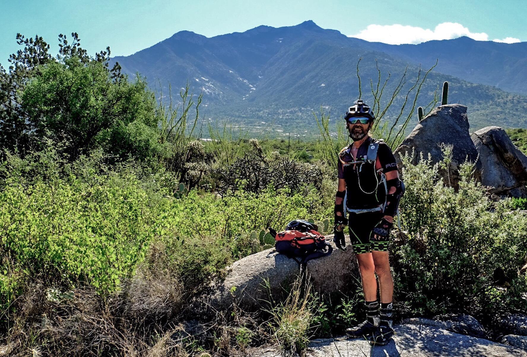 A person wearing mountain biking gear stands on a rock surrounded by greenery, with a backdrop of mountains under a clear blue sky. They have a helmet with a camera attached, gloves, and a hydration pack. A backpack rests on the ground nearby. 50-year Trail / Golder Ranch mountain bike trail.