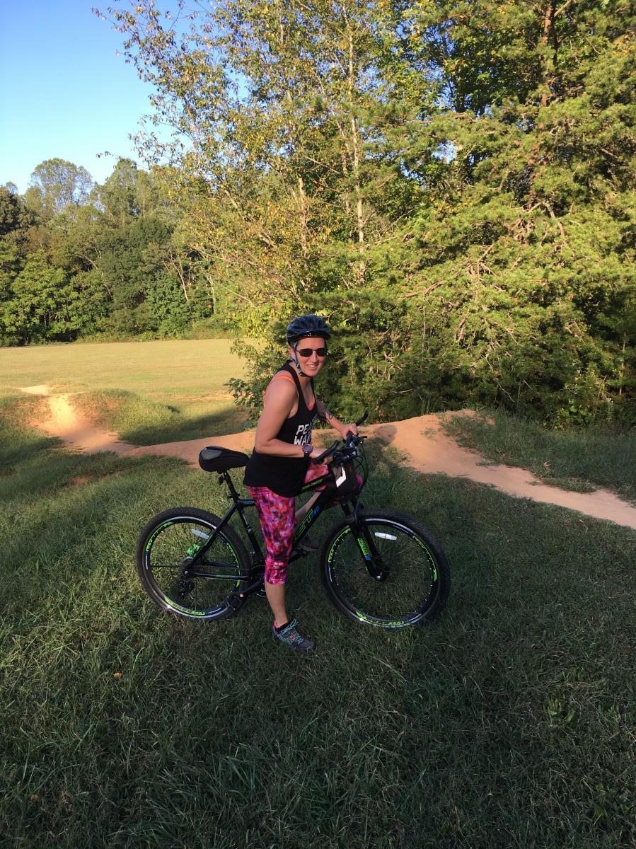 A person wearing a helmet and sunglasses stands next to a black bicycle on a grassy path surrounded by trees. The individual is dressed in a tank top and colorful leggings, smiling while holding the bike handlebar. Sunlight illuminates the scene, creating a vibrant outdoor atmosphere. Hobby Park mountain bike trail.